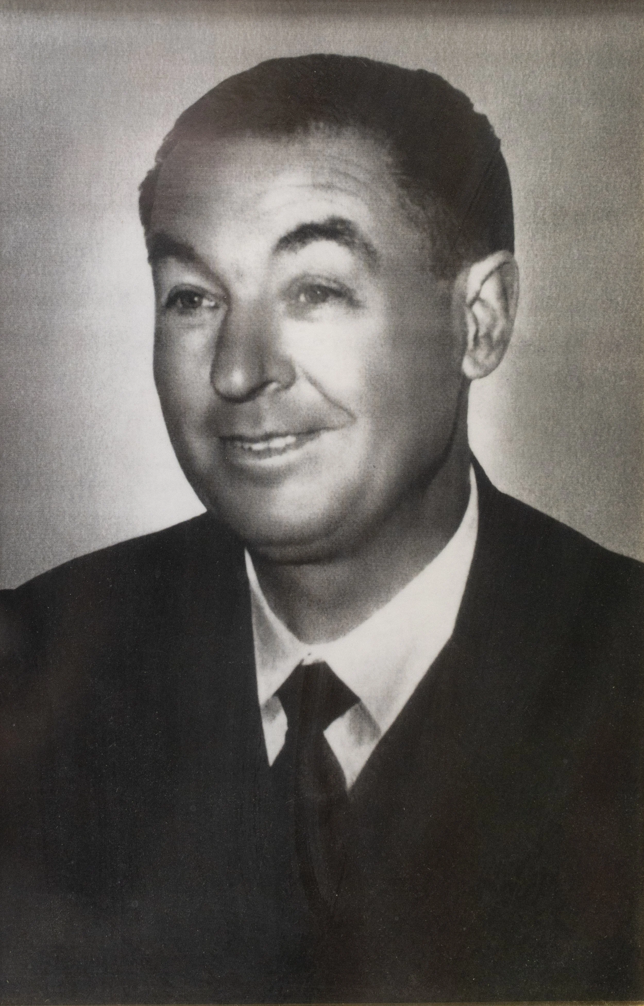 Retrato en blanco y negro de un hombre con cabello oscuro peinado hacia atrás, luciendo un traje formal con una camisa blanca y corbata, sonriendo ligeramente.