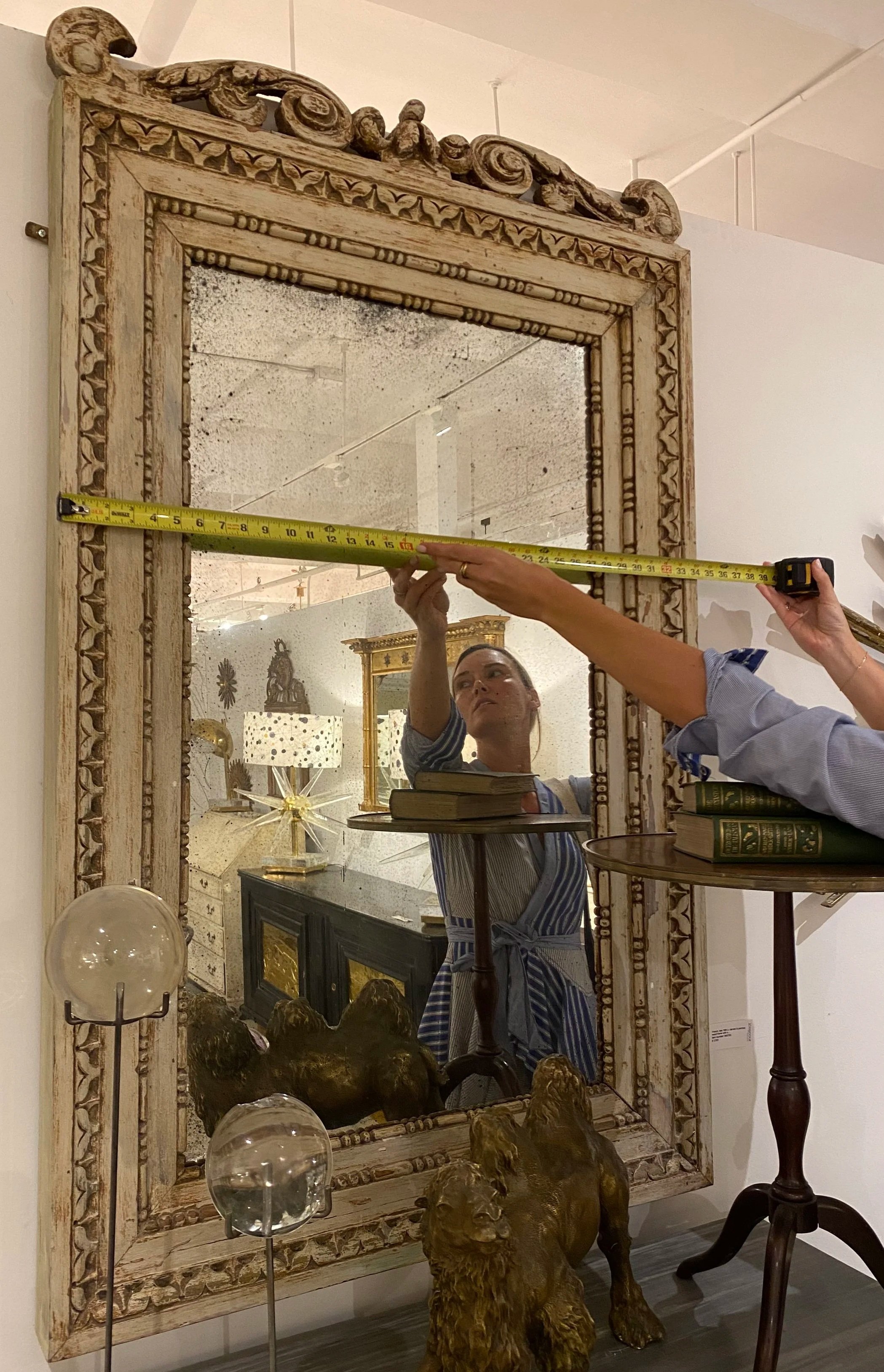 A mirror with an ornate carved wooden frame being measured with a tape measure by a woman, part of her reflection visible. The room contains decorative items, including a bronze lion sculpture, spherical glass objects, and various furniture pieces.