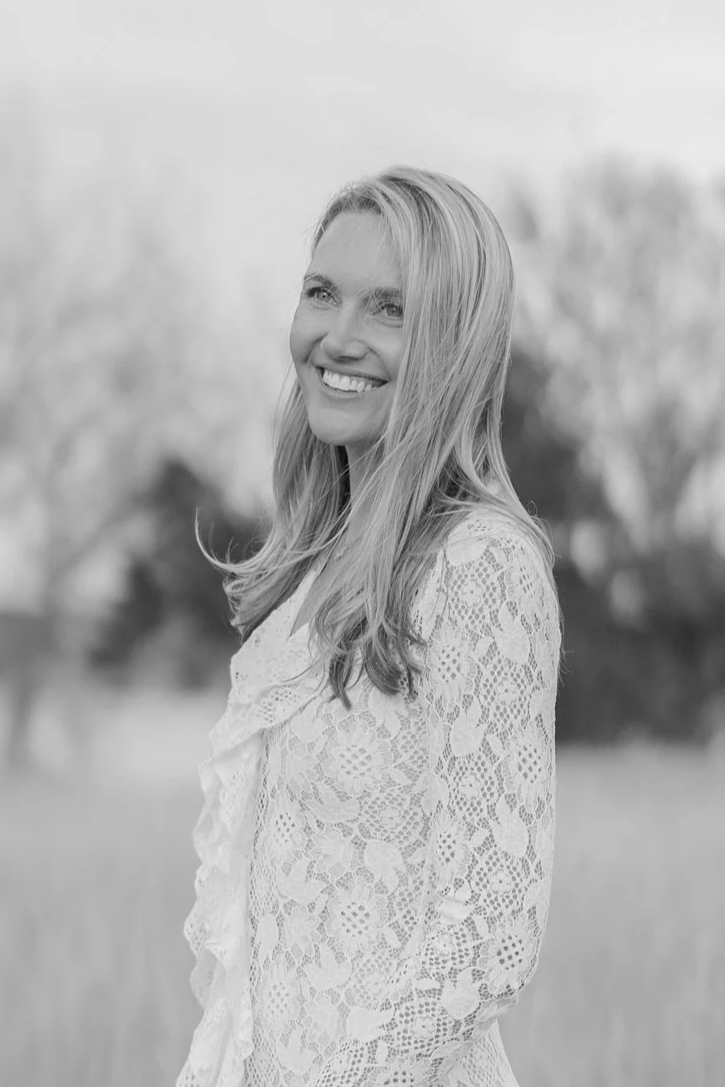 Black and white photo of a woman smiling, standing outdoors in a field, wearing a lace top.