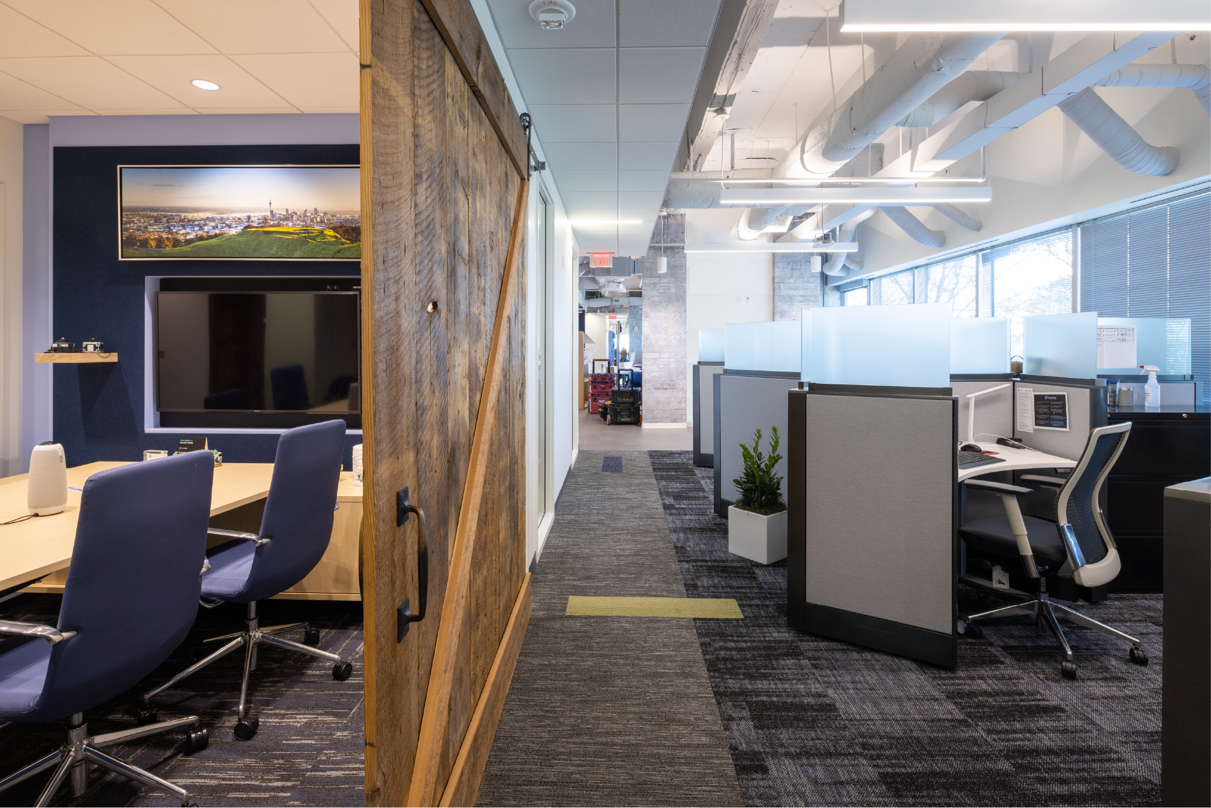 Open office space with cubicles, chairs, and a large monitor on a dark blue accent wall. There is a rustic wooden door or partition in the foreground. The ceiling has exposed white ducts and modern linear lighting. Large windows with blinds let in natural light.