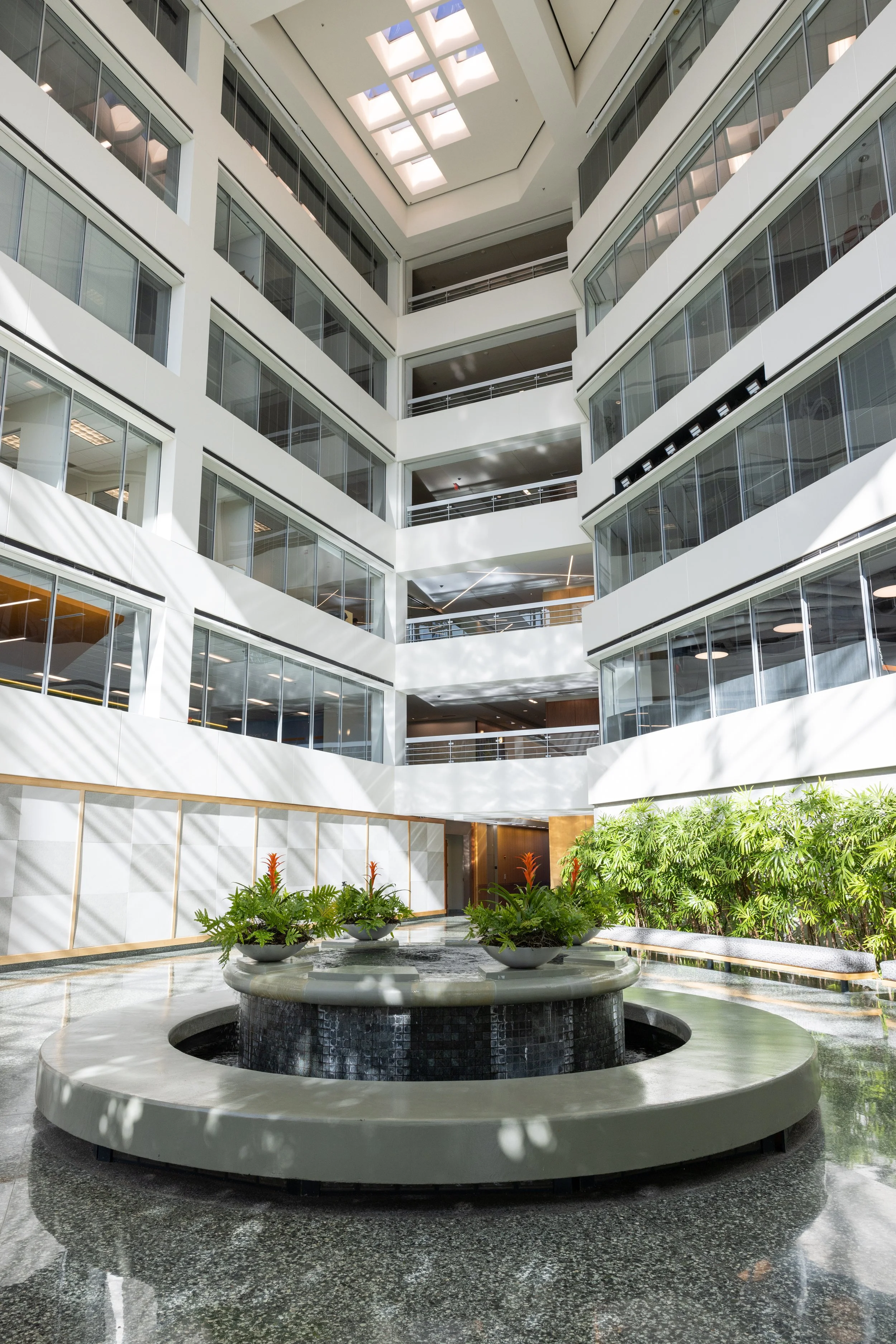 Interior view of a modern multi-story office building with glass walls, a large circular fountain with plants, and an open atrium with natural light from a skylight.