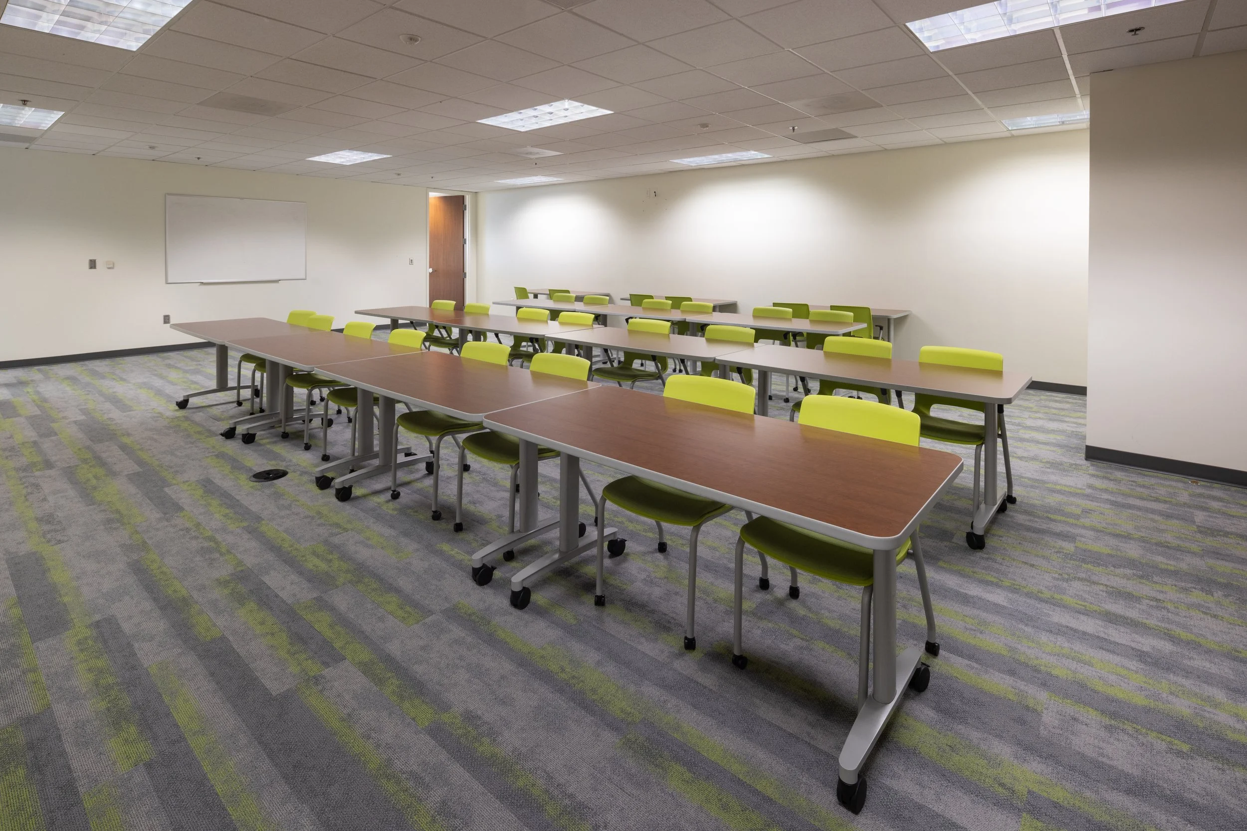 An empty classroom with long tables and lime green chairs on a patterned gray and green carpet, with a whiteboard on a white wall and a closed brown door.