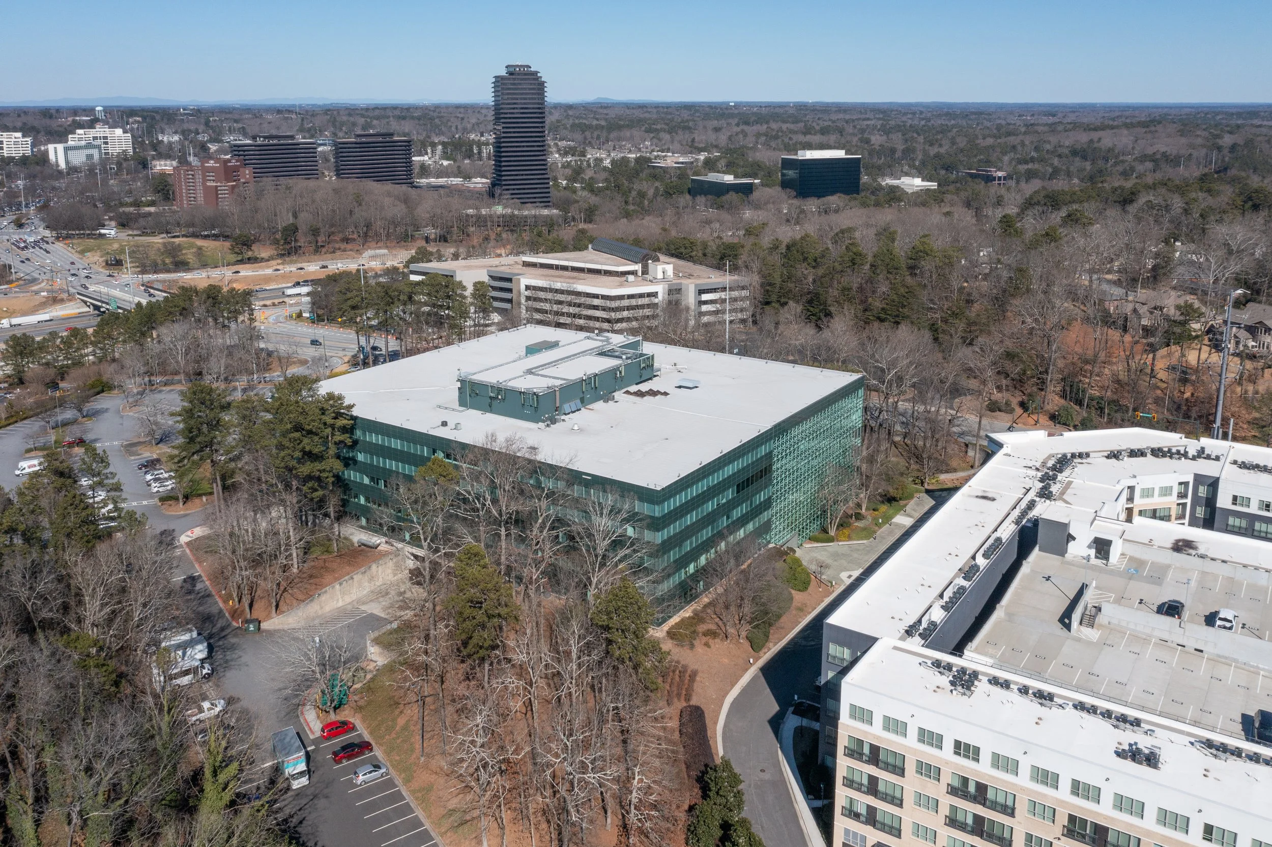 Aerial view of a cityscape with modern high-rise buildings, trees, a parking lot, and roads under a clear blue sky.