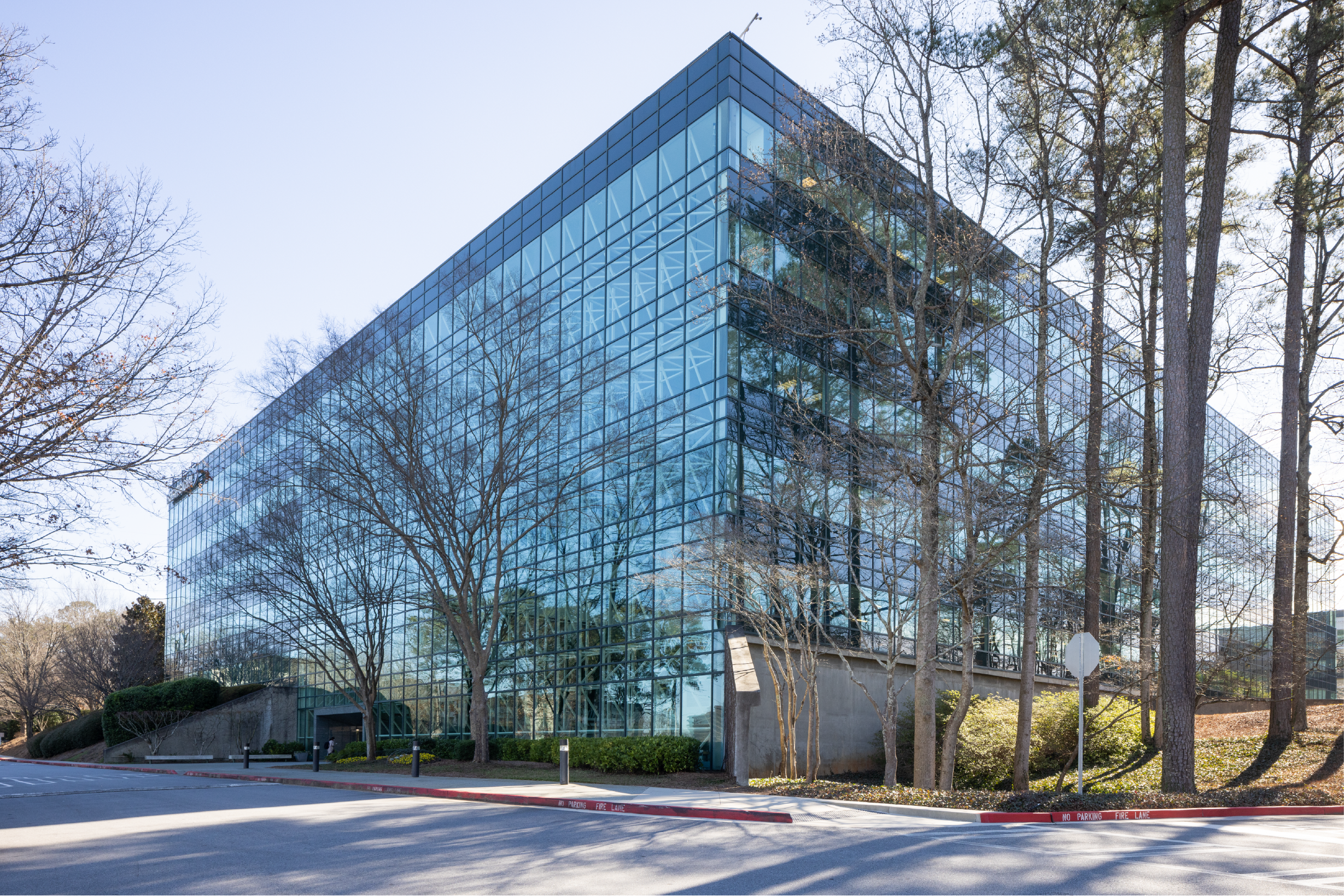 A modern glass office building with reflective windows surrounded by leafless trees, with a parking lot and no parking fire lane in the foreground.