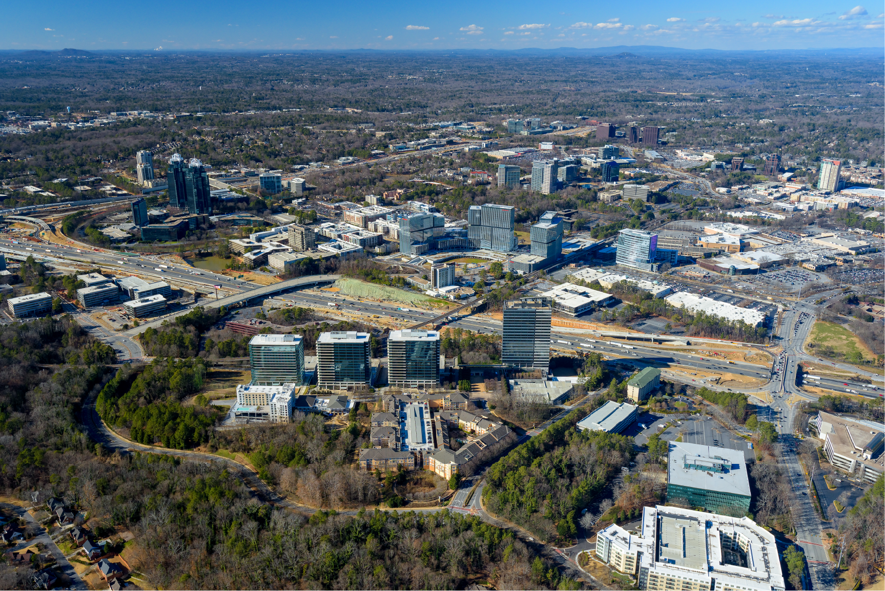 Aerial view of a cityscape with a mix of high-rise office buildings, residential areas, and highways with cars, surrounded by trees and open land under a partly cloudy sky.