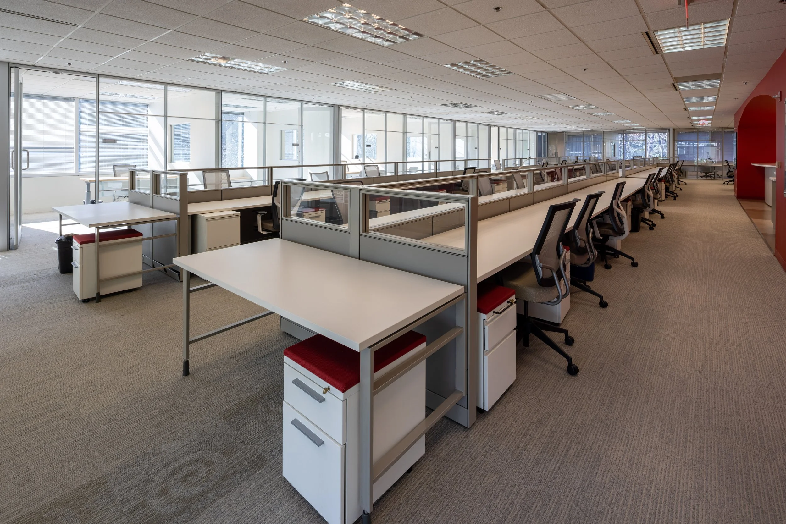 An empty office space with cubicles, desks, and chairs, featuring large floor-to-ceiling windows and a ceiling with fluorescent light panels.