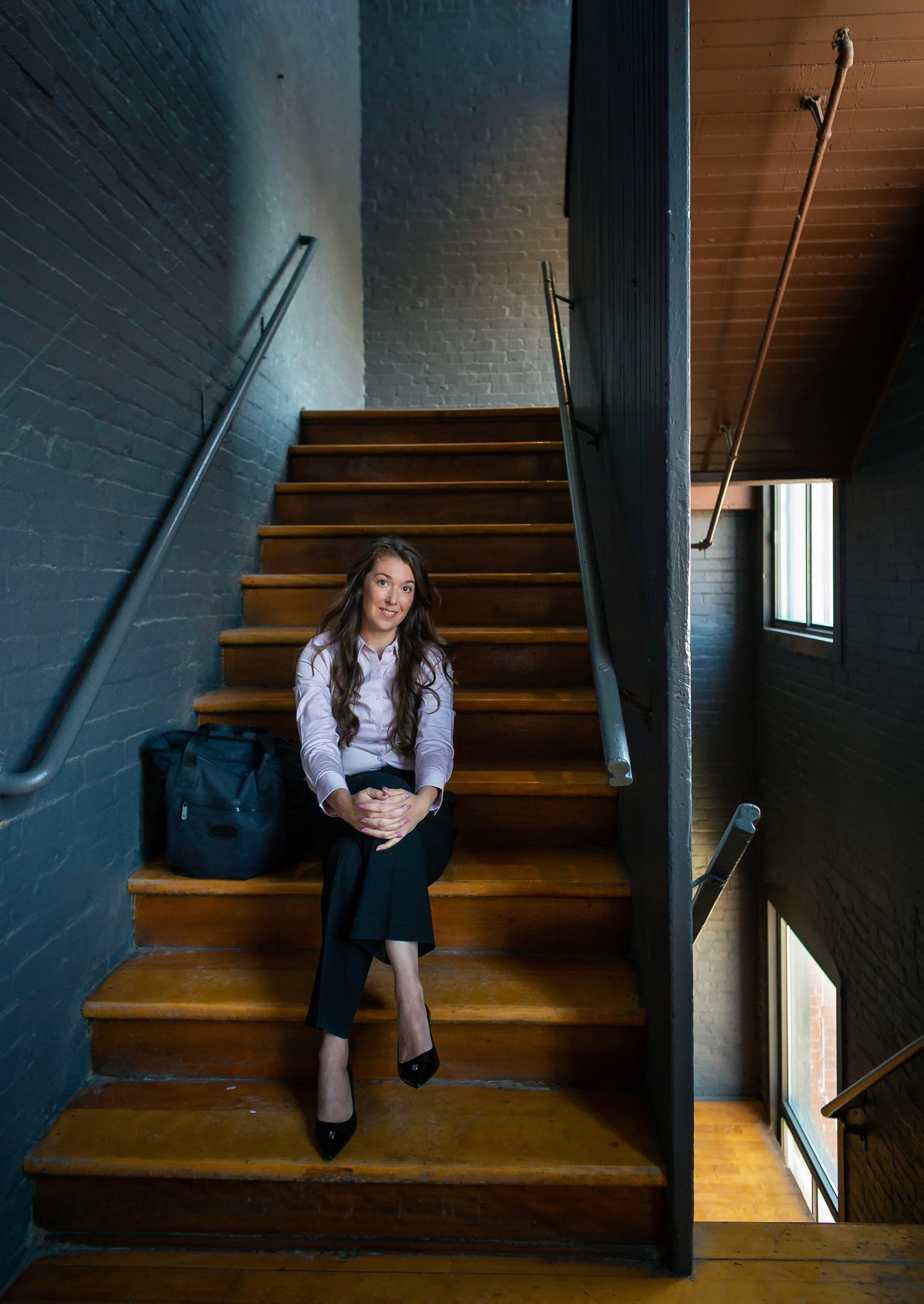 A woman sitting on wooden stairs inside a building, near a window. She has long brown hair, wears a white blouse, black pants, and black heels. There is a black backpack next to her.