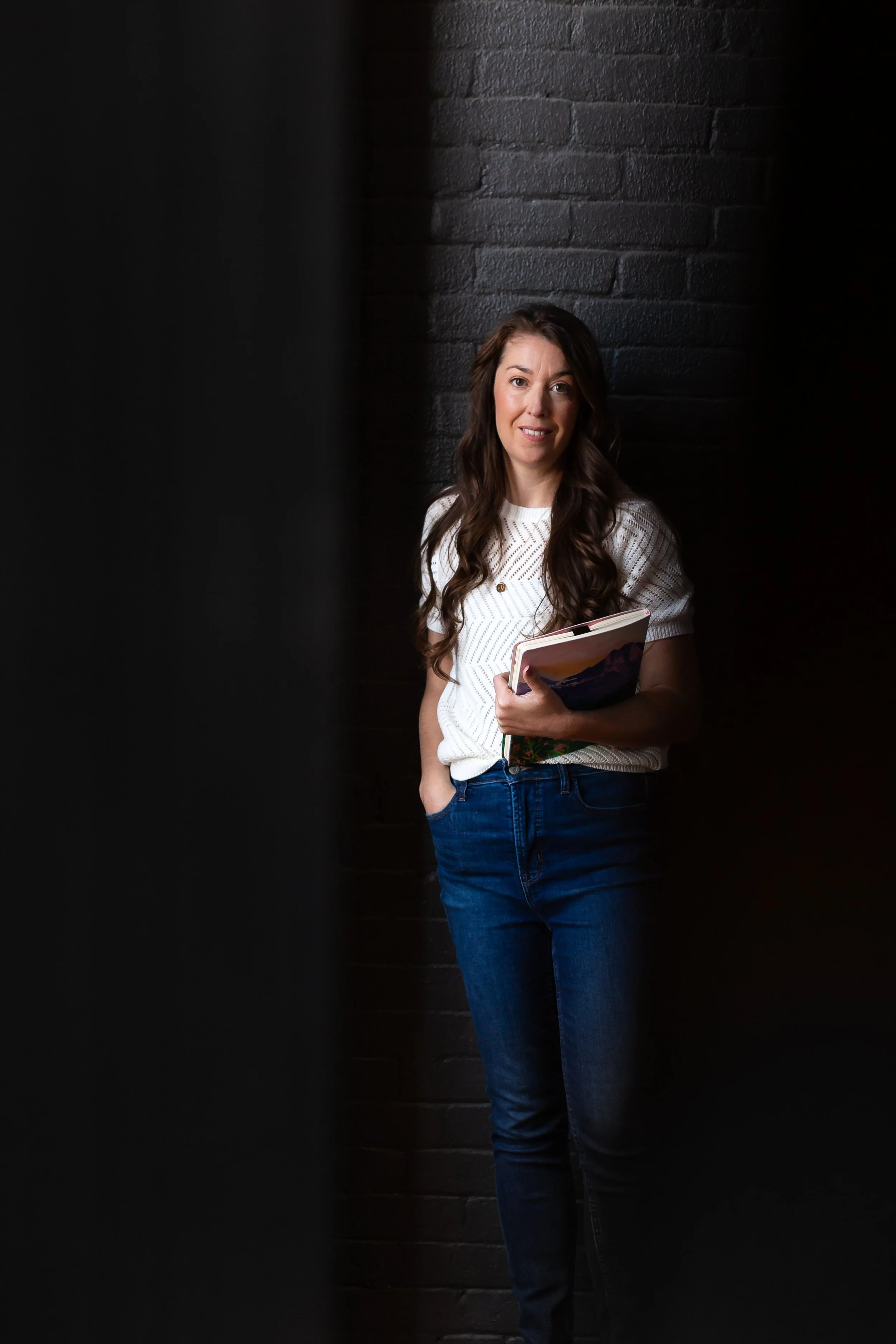 A woman holding notebooks, standing against a dark brick wall, with partial dark framing in the foreground.