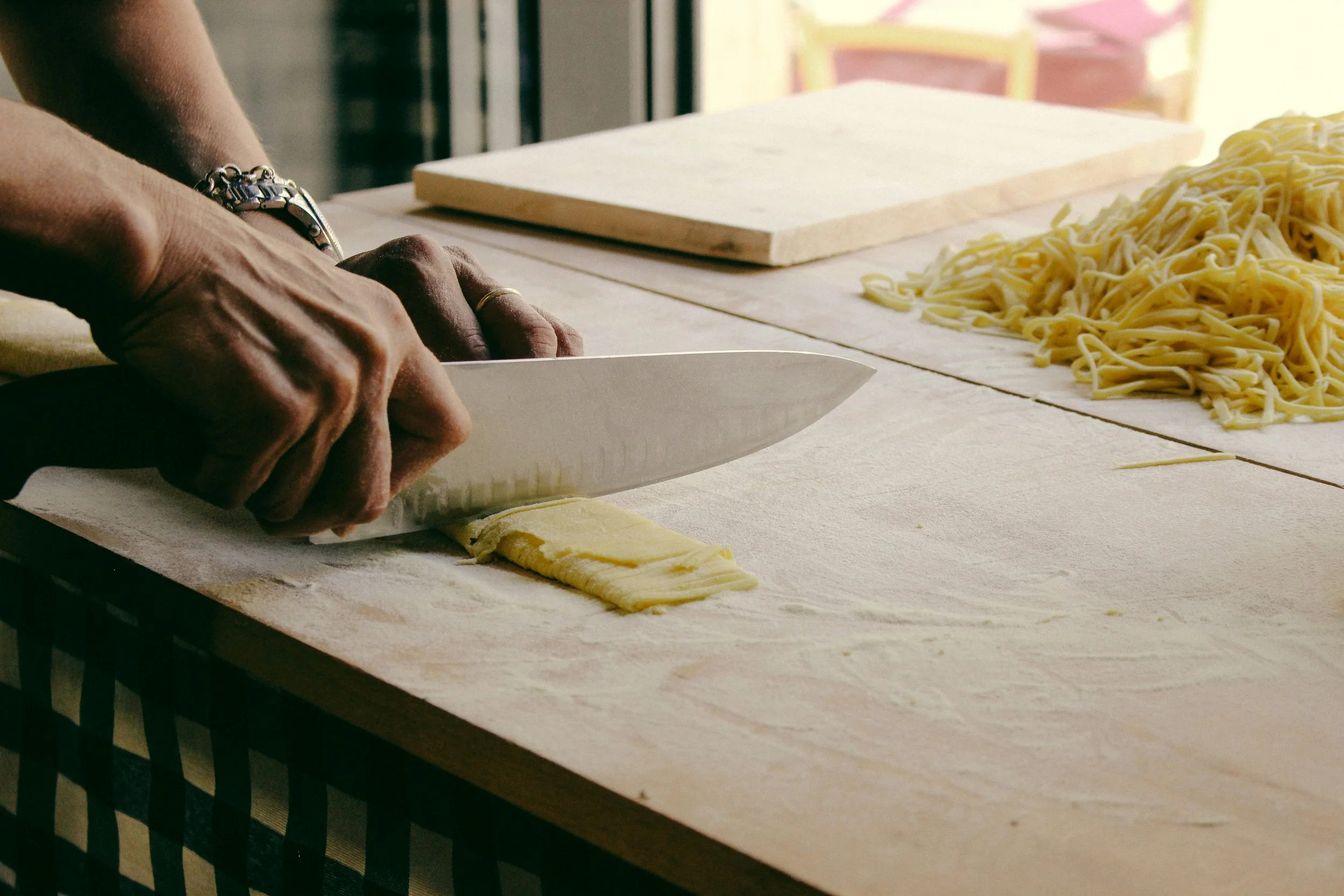 Person schneidet Teig auf einer Holzarbeitsfläche, neben einem Stapel frisch gekochter Pasta.