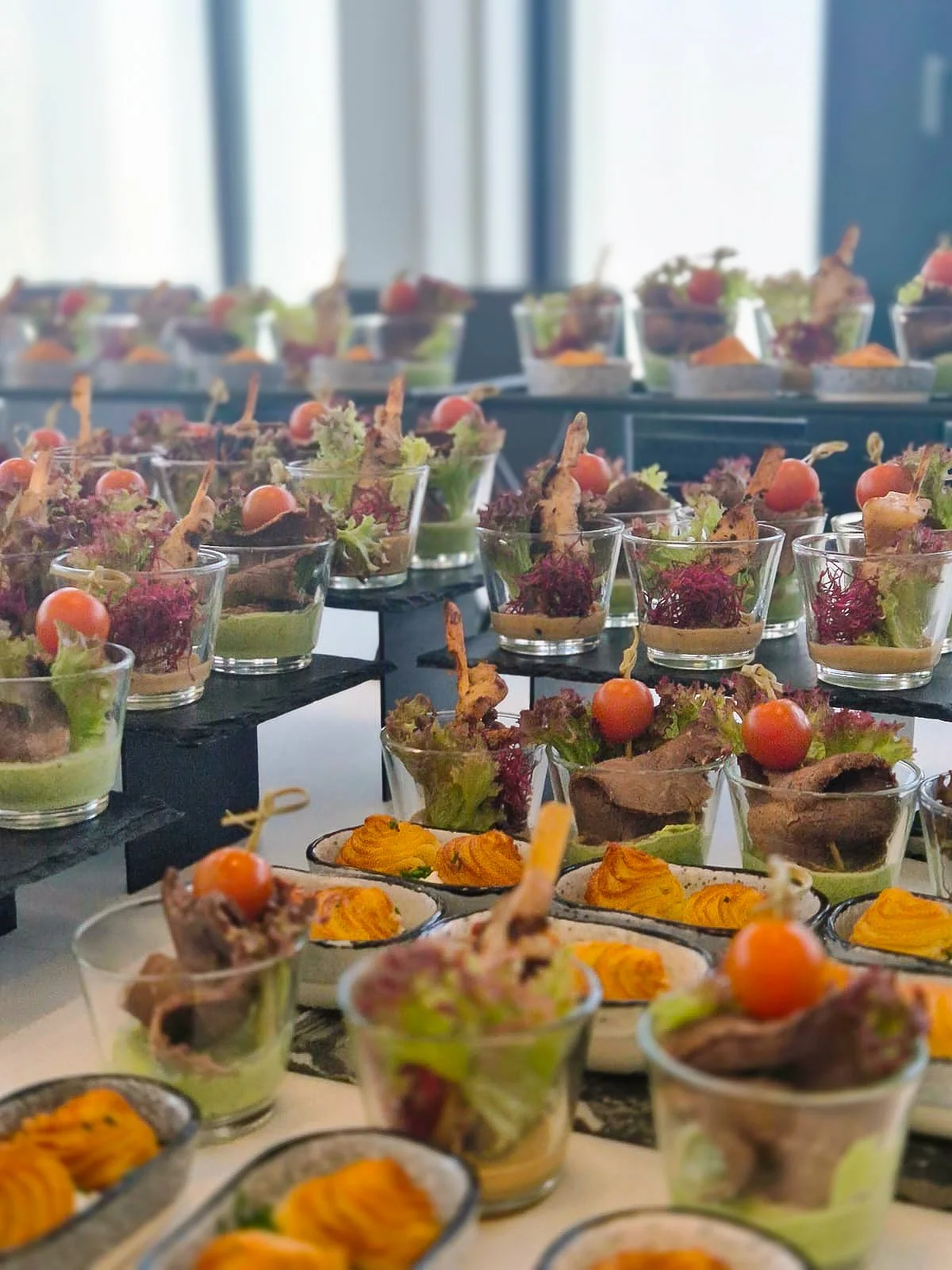 Verschiedene Canapés mit Salat, Tomate und Fleisch in Gläsern, dekorativ serviert auf schwarzen und weißen Platten.