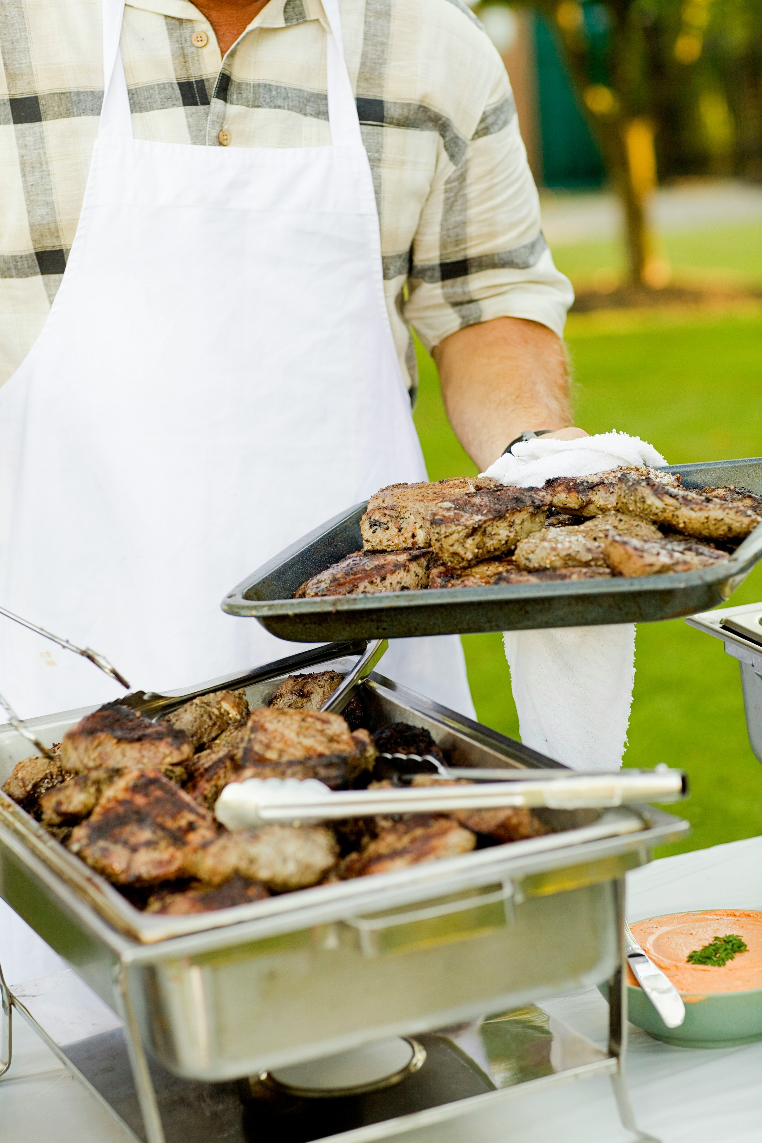 Ein Mann in kariertem Hemd und weißer Schürze serviert gegrillte Fleischstücke auf einem Buffet im Freien.