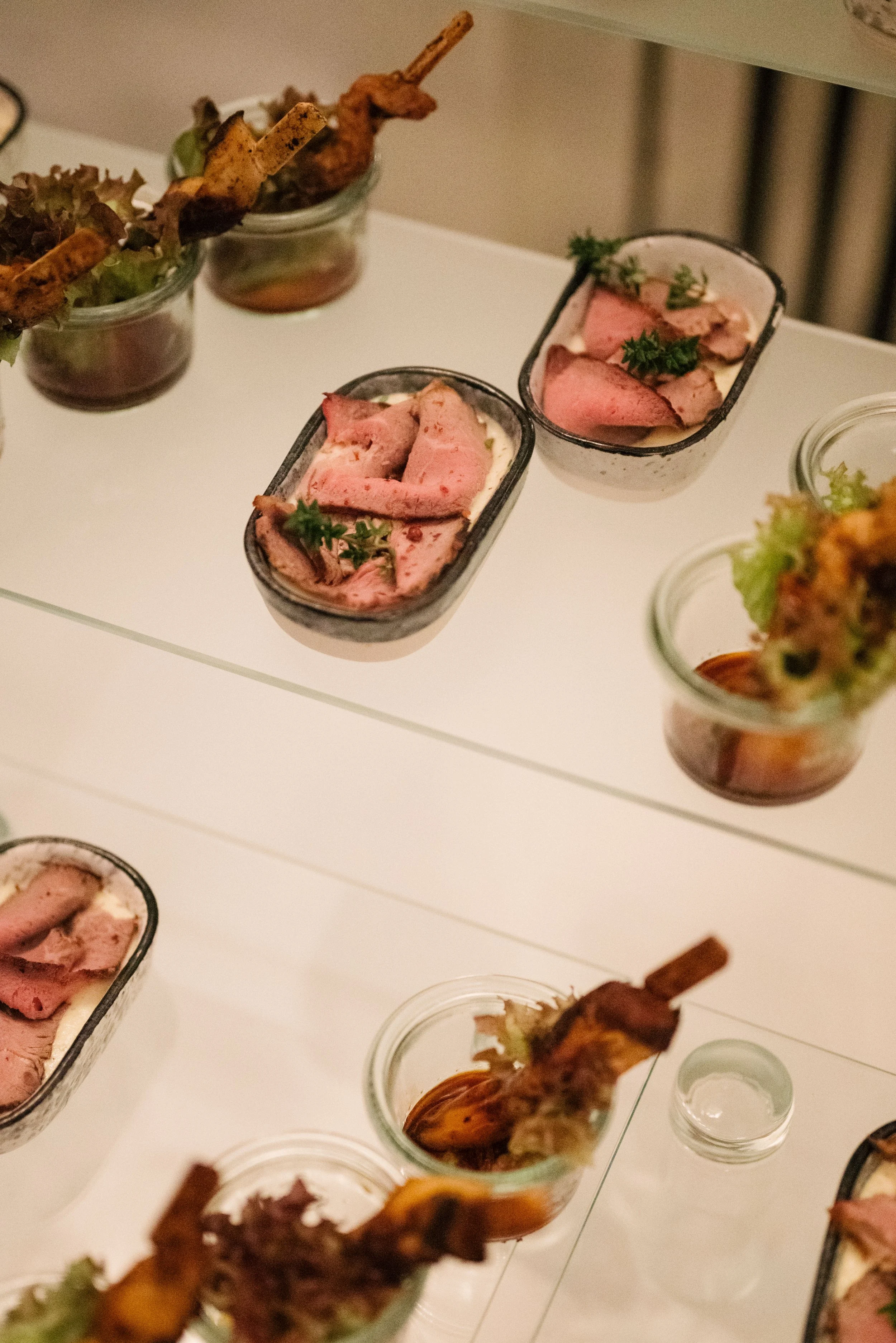 Verschiedene Häppchen in kleinen Gläsern auf einem weißen Tisch, darunter Roastbeef mit Kräutern und marinierte Gemüse.