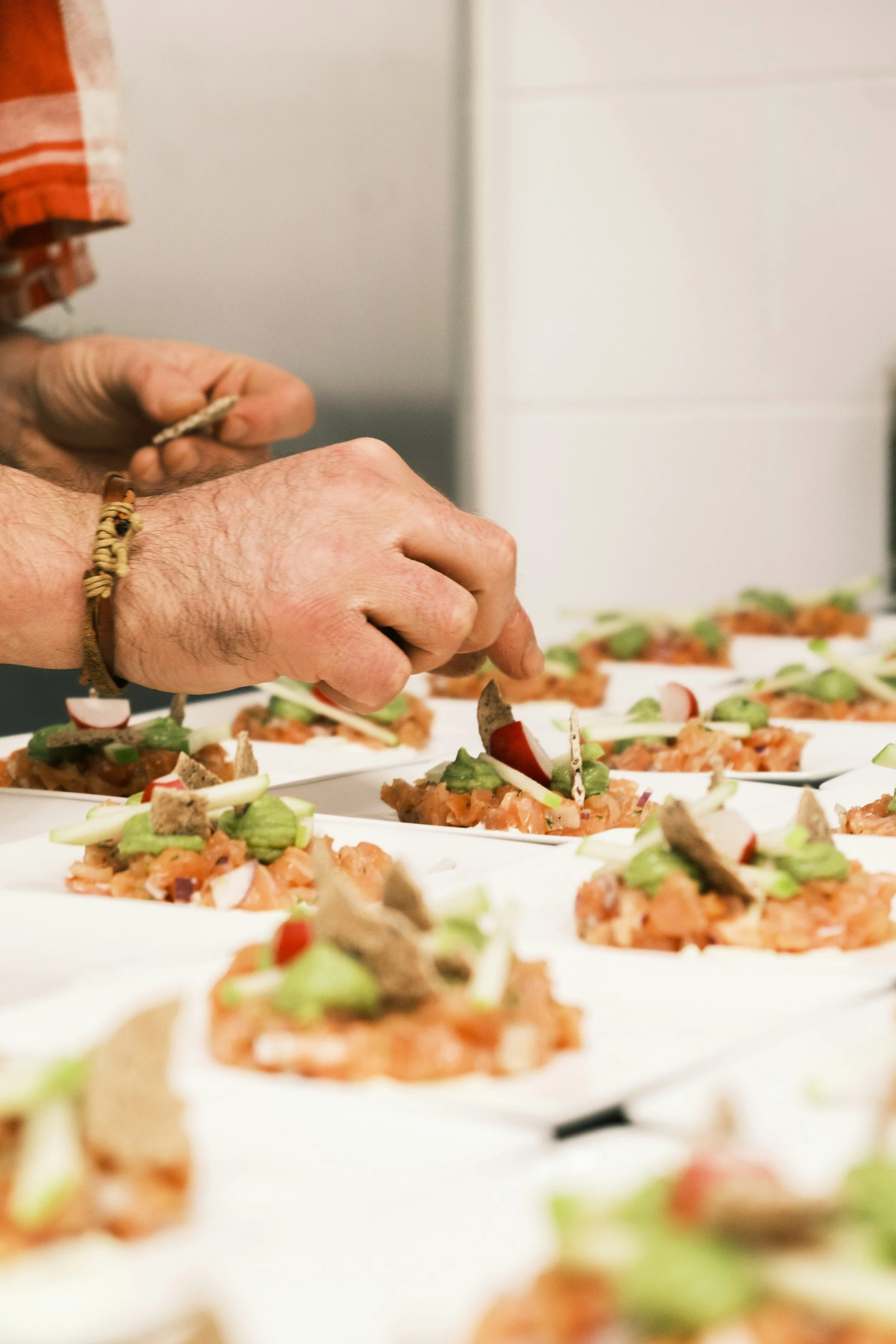 Nahaufnahme von einer Person, die kleine Snacks oder Bruschetta mit Avocado, Radieschen, Tomaten und Brot auf weißen Tellern zubereitet.