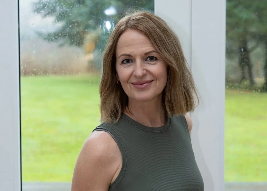 Close-up of a woman with shoulder-length light brown hair, smiling, wearing a sleeveless gray top, standing indoors near a large window with a blurred outdoor garden in background.
