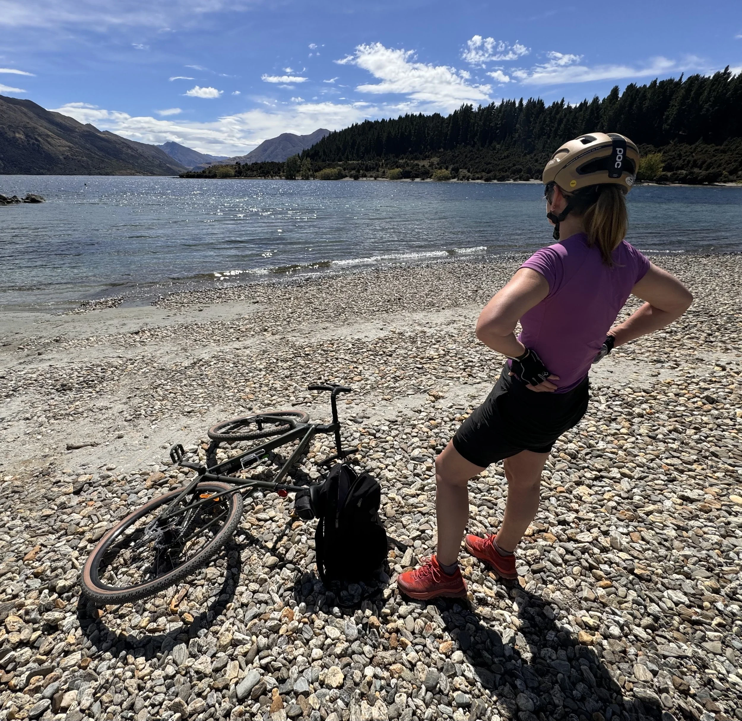 A person wearing a helmet and athletic clothing sitting on a rocky lakeshore with a mountain range and a forested hill in the background under a partly cloudy sky.