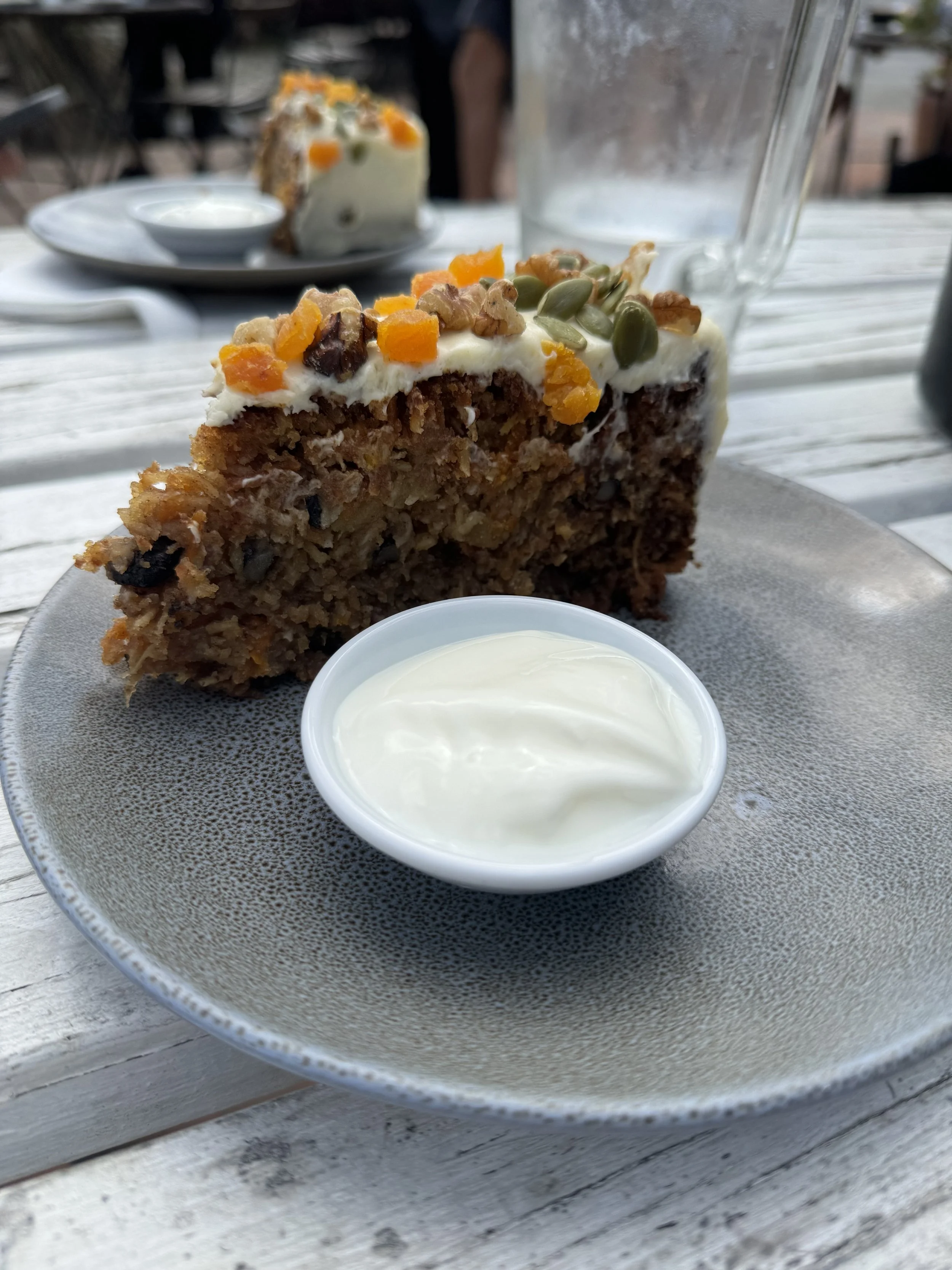 Close-up of a slice of carrot cake with cream cheese frosting topped with nuts, pumpkin seeds, and dried fruit on a gray plate with a small bowl of white frosting or cream, on a wooden table outside.