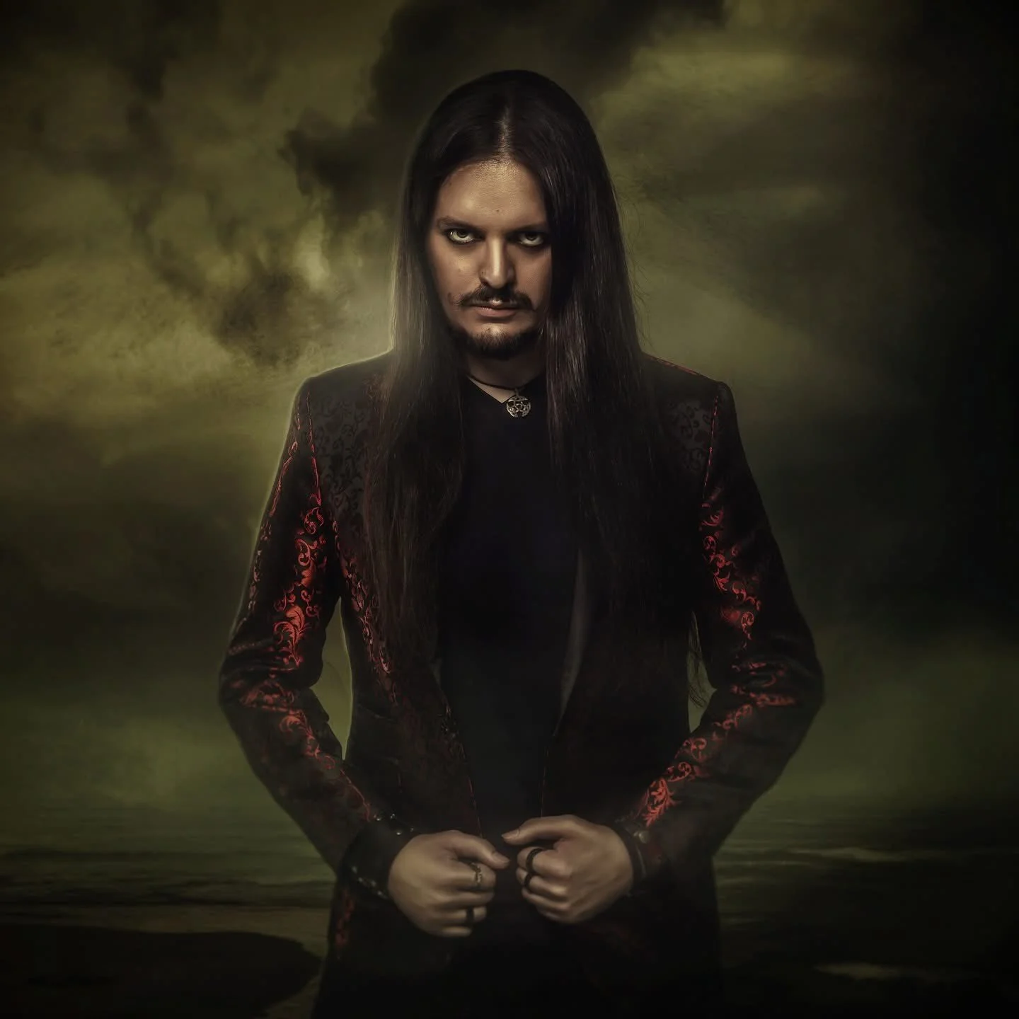 A man with long dark hair, wearing a black and red embroidered blazer, standing with hands clasped in front, against a dark, cloudy background resembling a stormy sky.