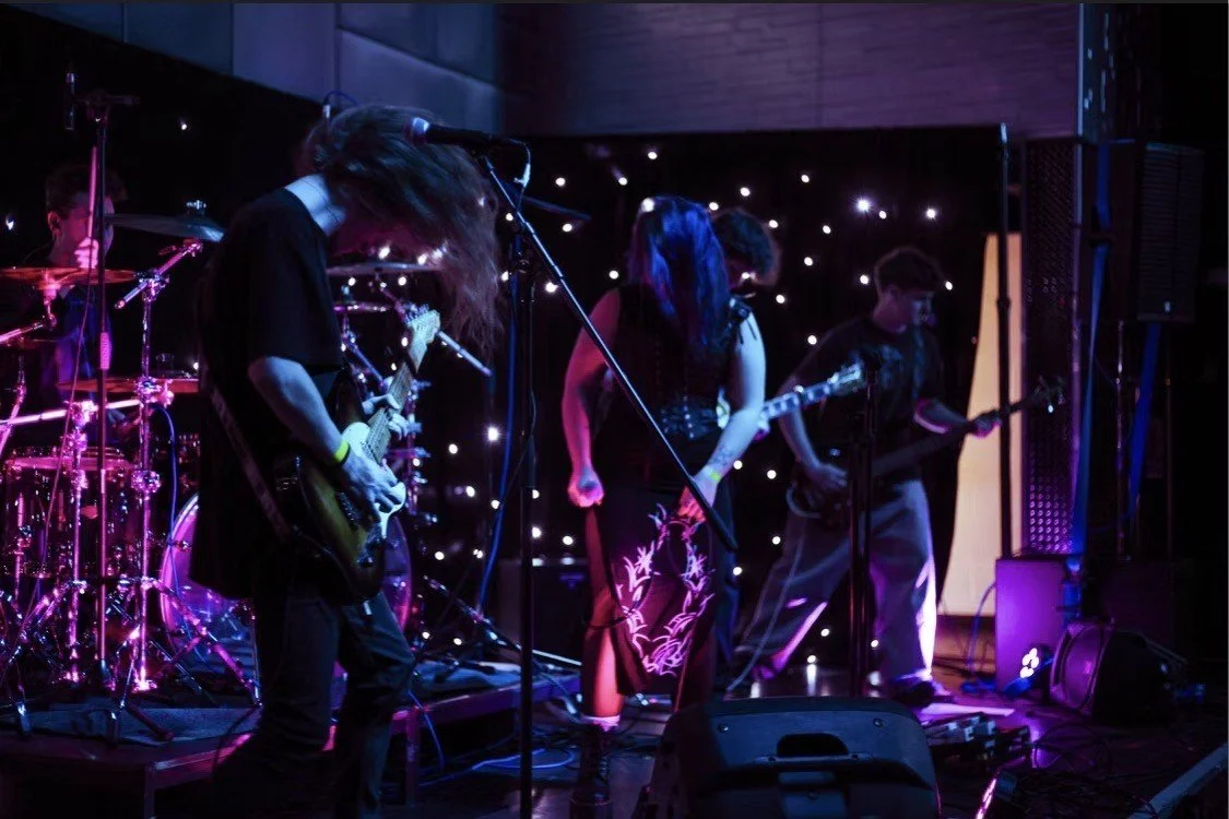 A band performing on stage with purple and blue lighting, featuring a drummer, two guitarists, and a female vocalist.