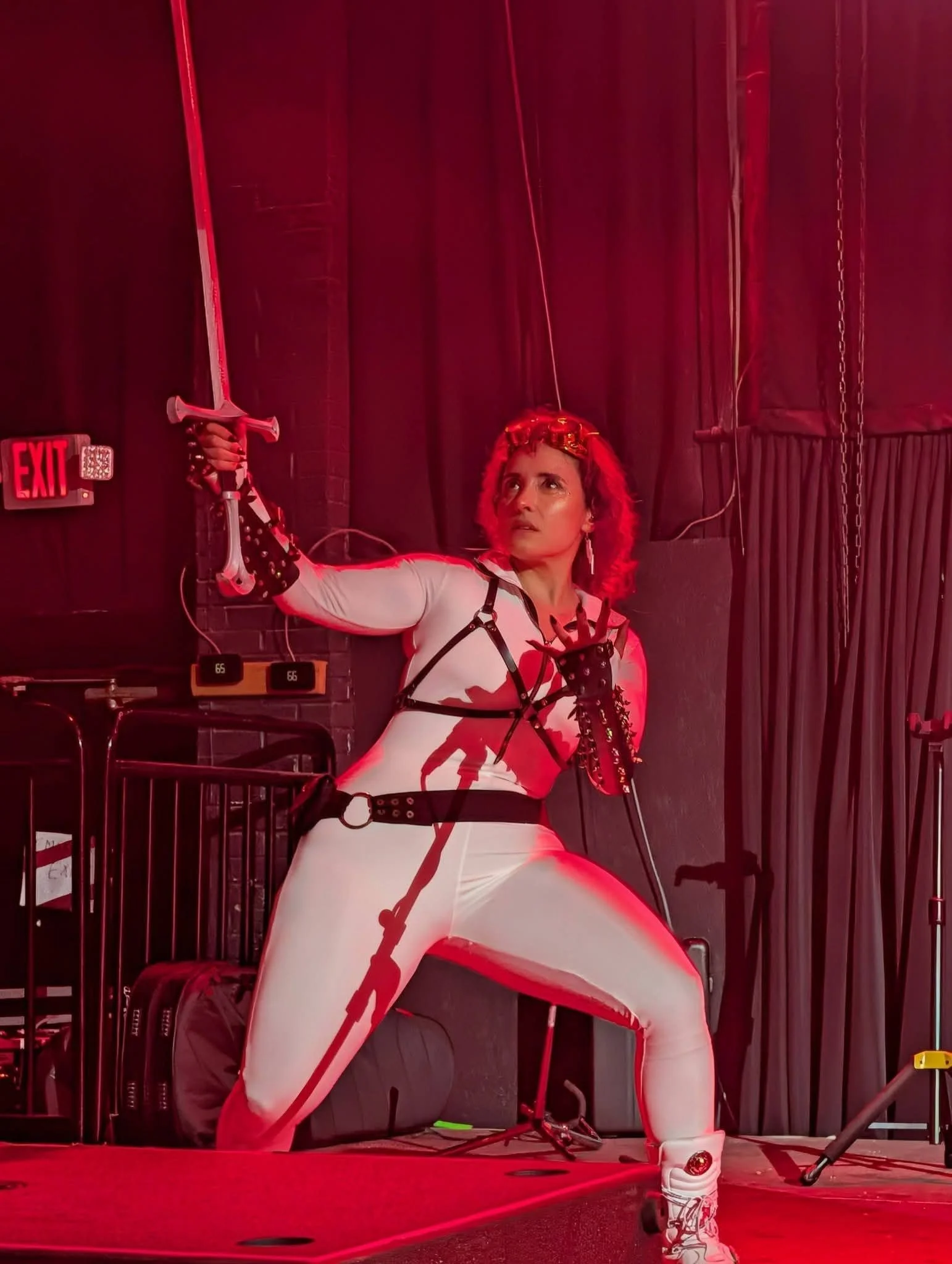 A woman dressed in futuristic white and black clothing with red accents is kneeling on stage with one arm extended holding a sword, in a dimly lit setting illuminated by red stage lights.