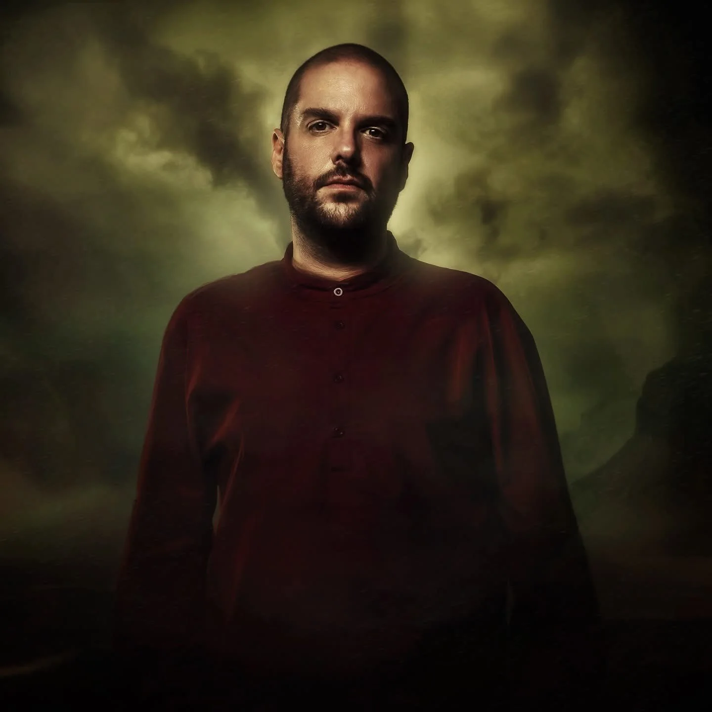 A man with a beard and shaved head wearing a dark red shirt, standing against a cloudy yellow-green background.