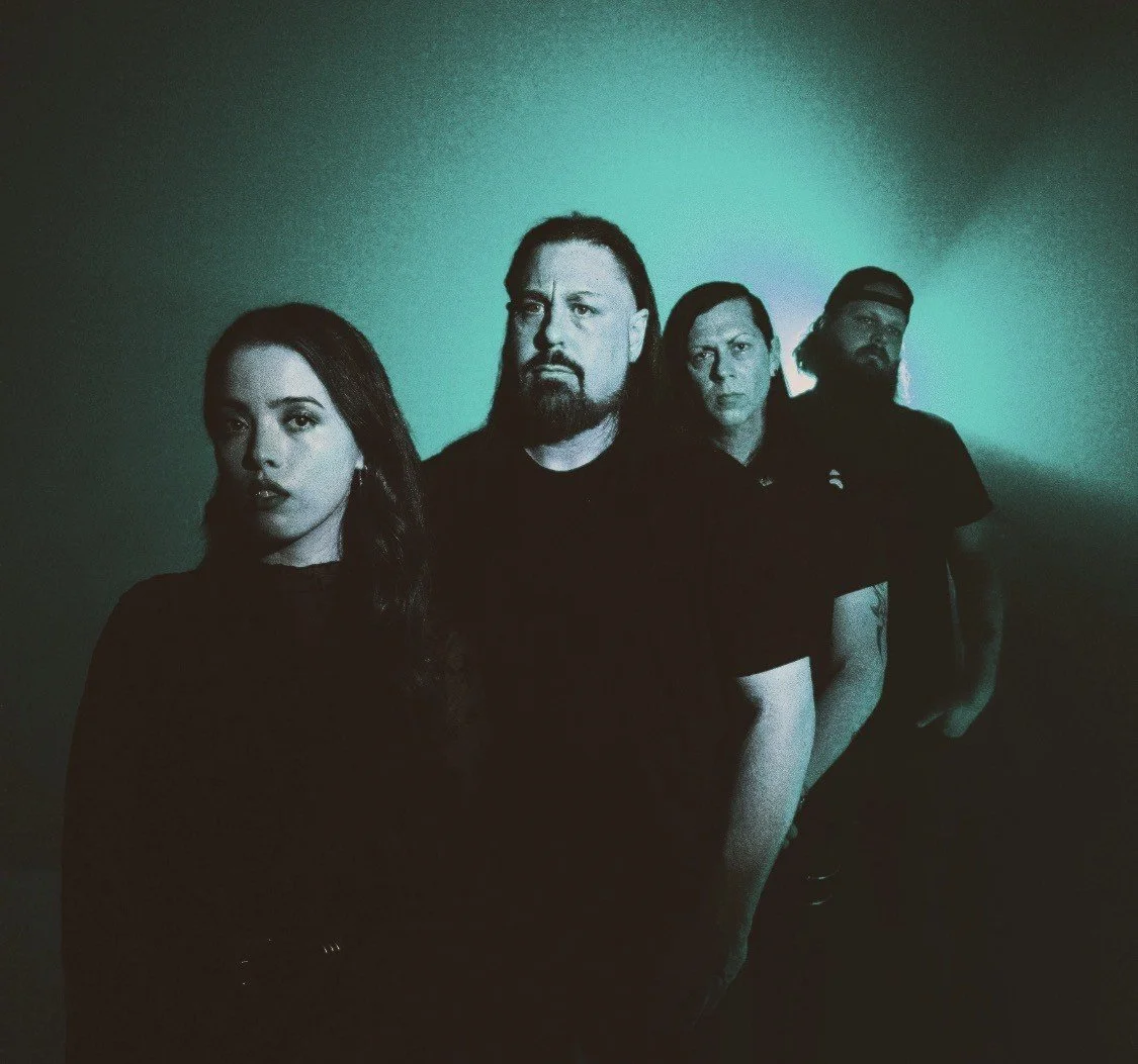 A group of four people standing in a line, with dramatic lighting, appearing serious, against a dark background.