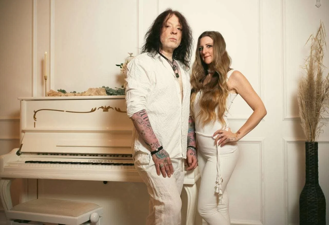 A man with long black hair, tattoos, and wearing white clothing stands next to a woman with long wavy brown hair in a white outfit, in front of a white piano and cream-colored wall decorated with candles and dried plants.