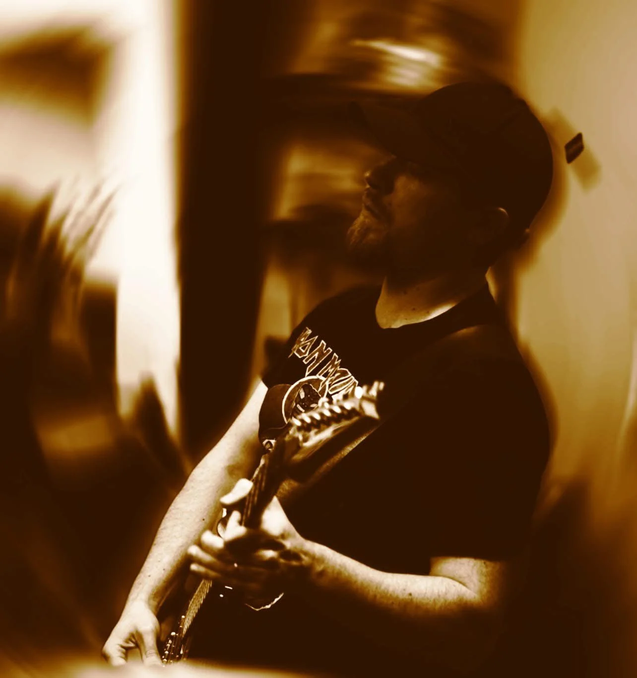 A person playing an electric guitar, wearing a baseball cap and a black t-shirt, in a room with warm lighting.