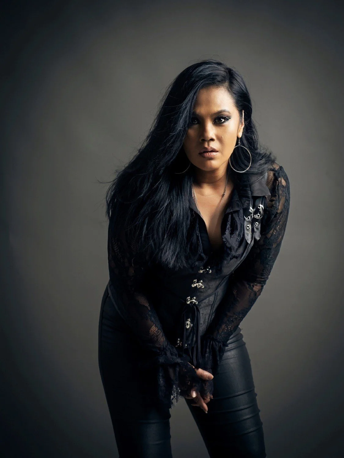 A woman with long black hair wearing a black lace top, black leather pants, and hoop earrings, posing against a gray background.