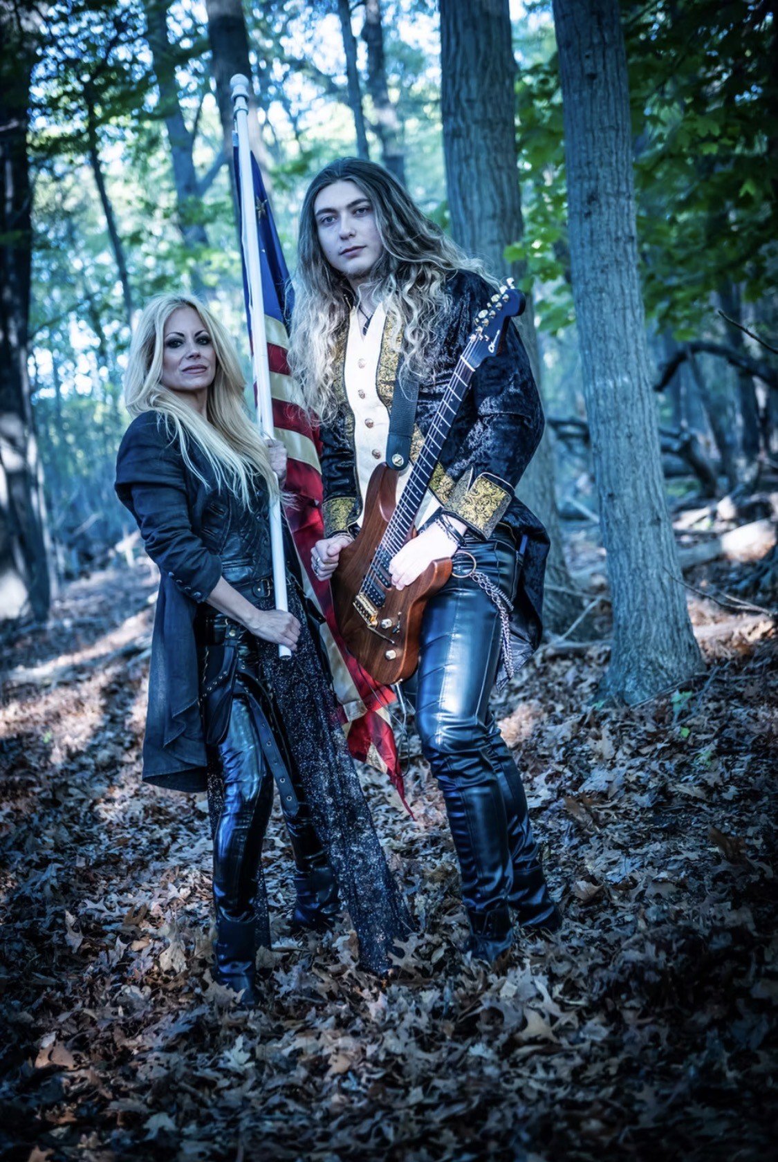 Two women standing in a wooded area, one holding an American flag and the other holding a guitar, dressed in rock-style clothing.