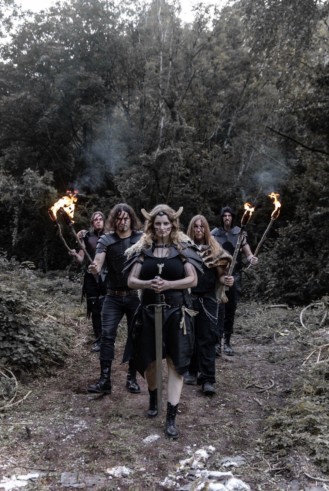 A group of five people dressed as fantasy warriors or Vikings walking through a wooded forest, carrying torches and weapons, with a woman wearing a horned helmet at the front.