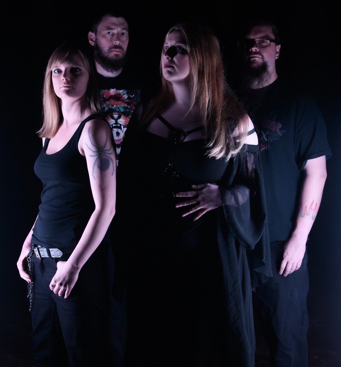 Four individuals standing against a black background, dressed in dark clothing with a gothic or alternative style, posing for a photo.
