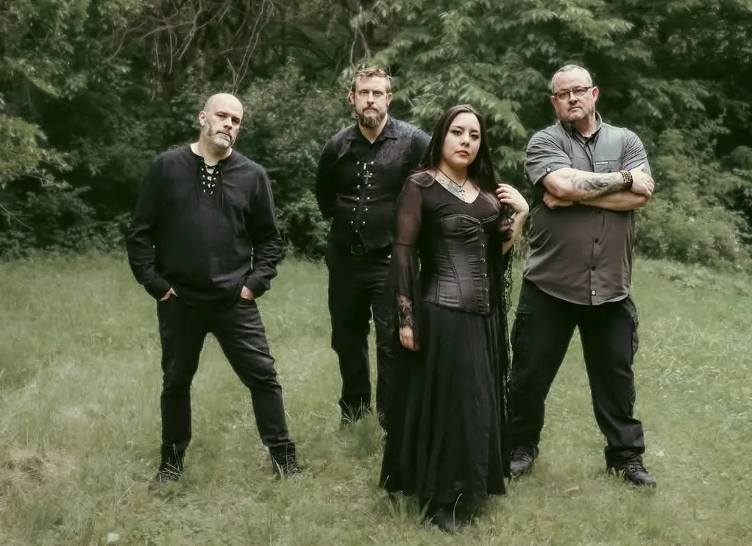 A group of four people standing outdoors on grass with trees in the background, dressed in dark, gothic-style clothing.
