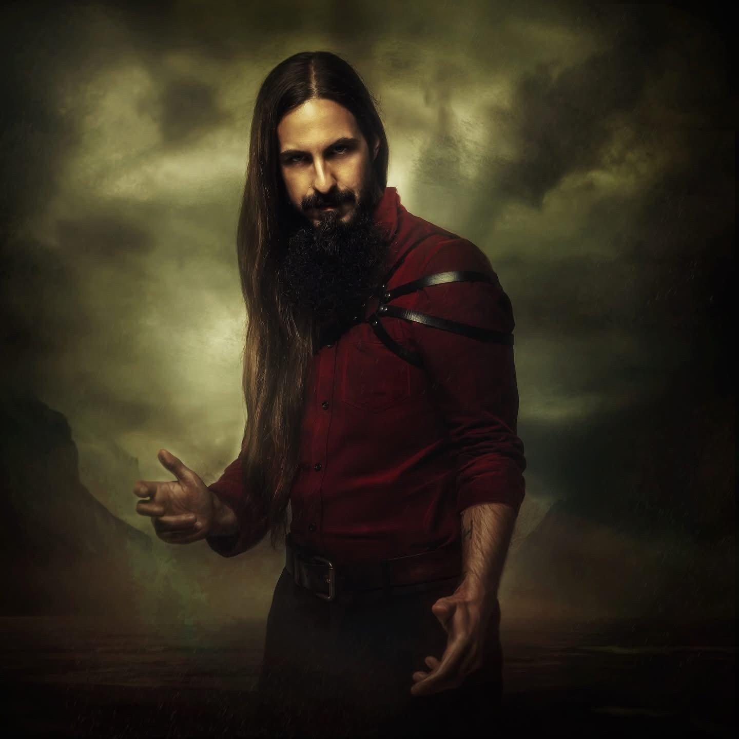 A man with long hair and a beard wearing a red shirt and black pants, standing against a dark, cloudy background, making a gesture with his right hand.
