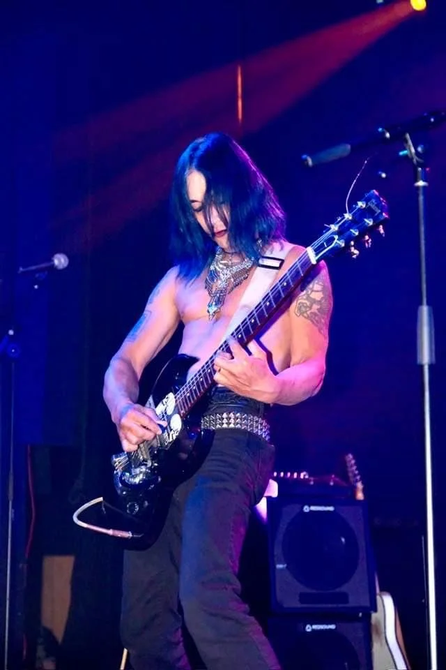 Female musician with black hair playing an electric guitar on stage with colorful lighting.