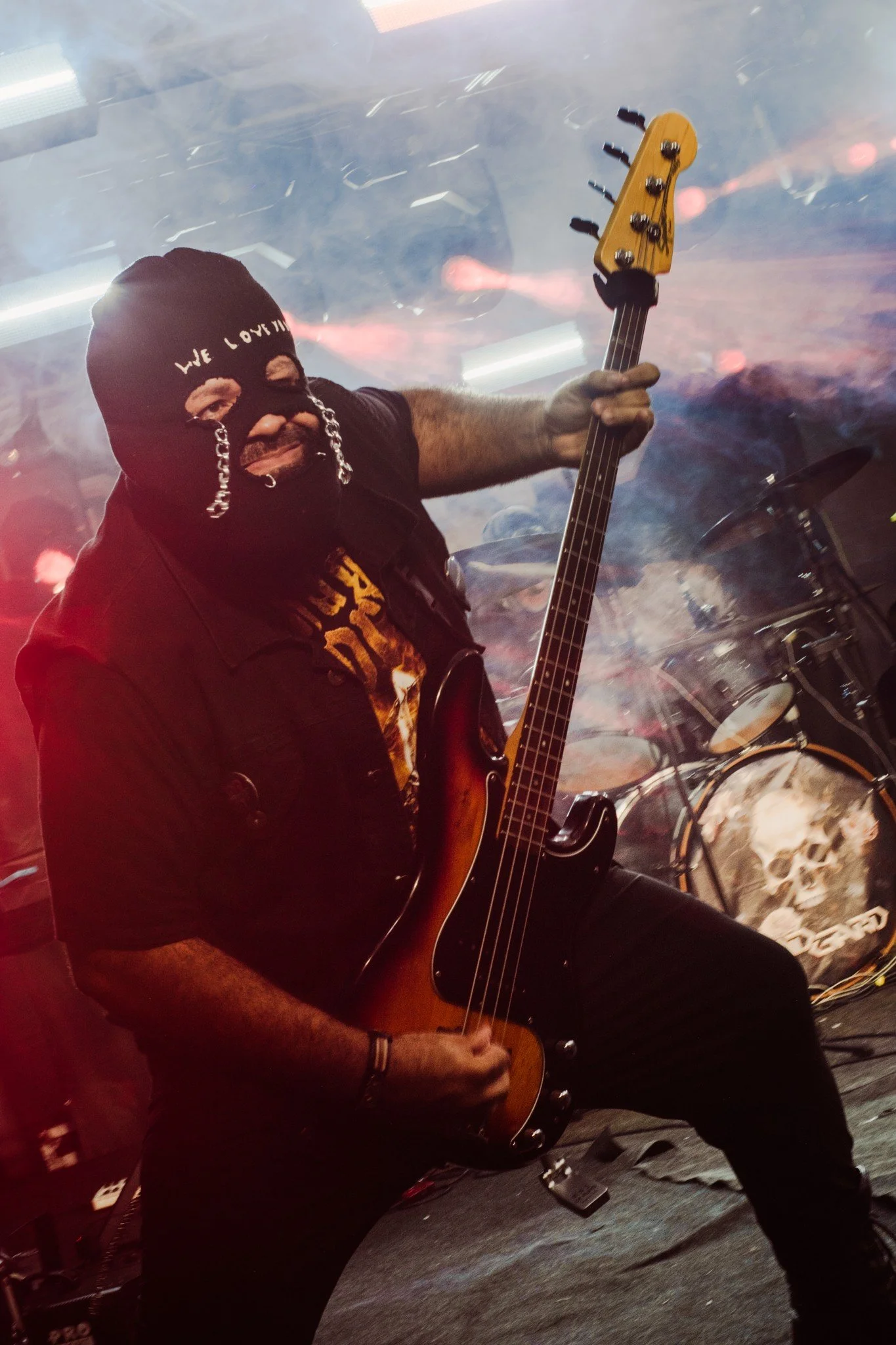 A man wearing a black ski mask and black gloves playing an acoustic guitar on stage. The background has a smoky, colorful atmosphere with some equipment visible.