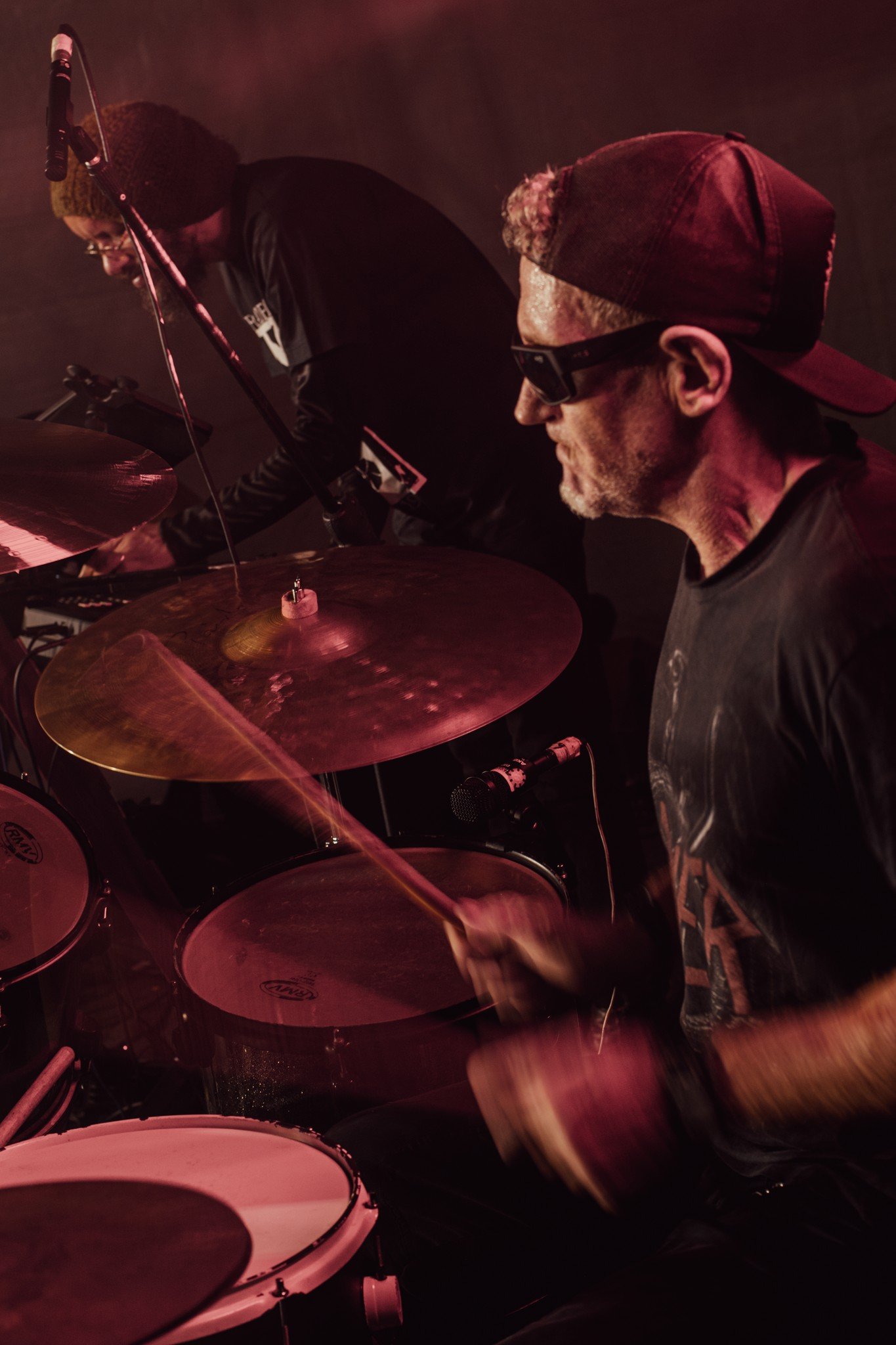 A man wearing a red cap backwards, sunglasses, and a black T-shirt playing drums on stage with another musician in the background, dimly lit with red lighting.