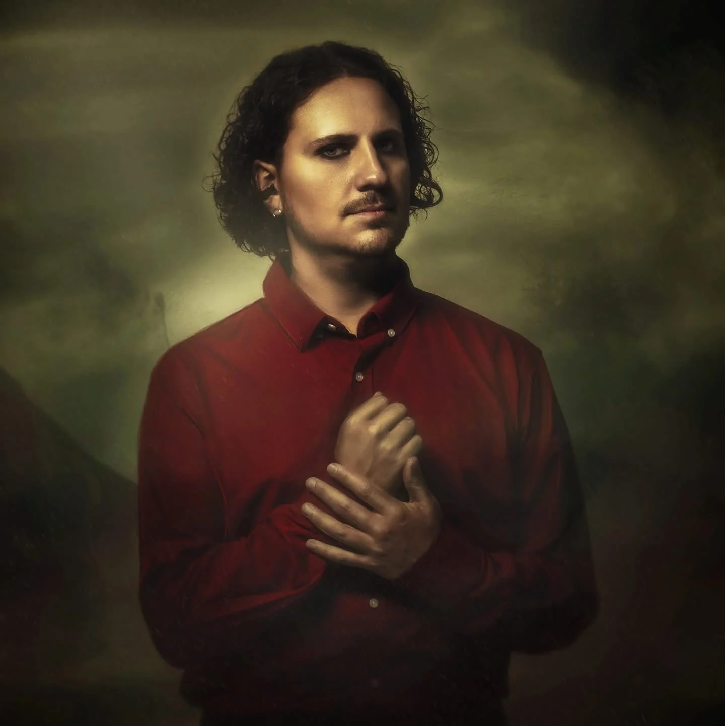 A man with curly dark hair, wearing a red shirt, standing against a dark, muted background, with one hand holding the collar of his shirt and the other crossed over his chest.