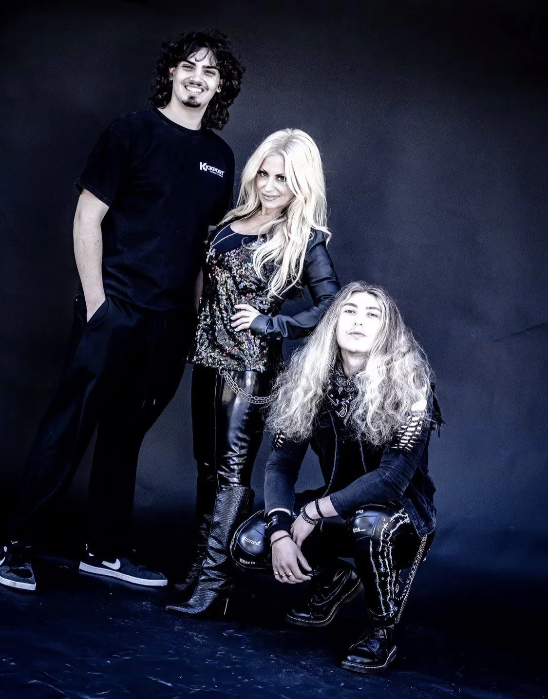 Three musicians posing in front of a dark background, a man standing on the left with curly hair, a woman with long platinum blonde hair in the middle, and a woman with voluminous curly hair kneeling in front.