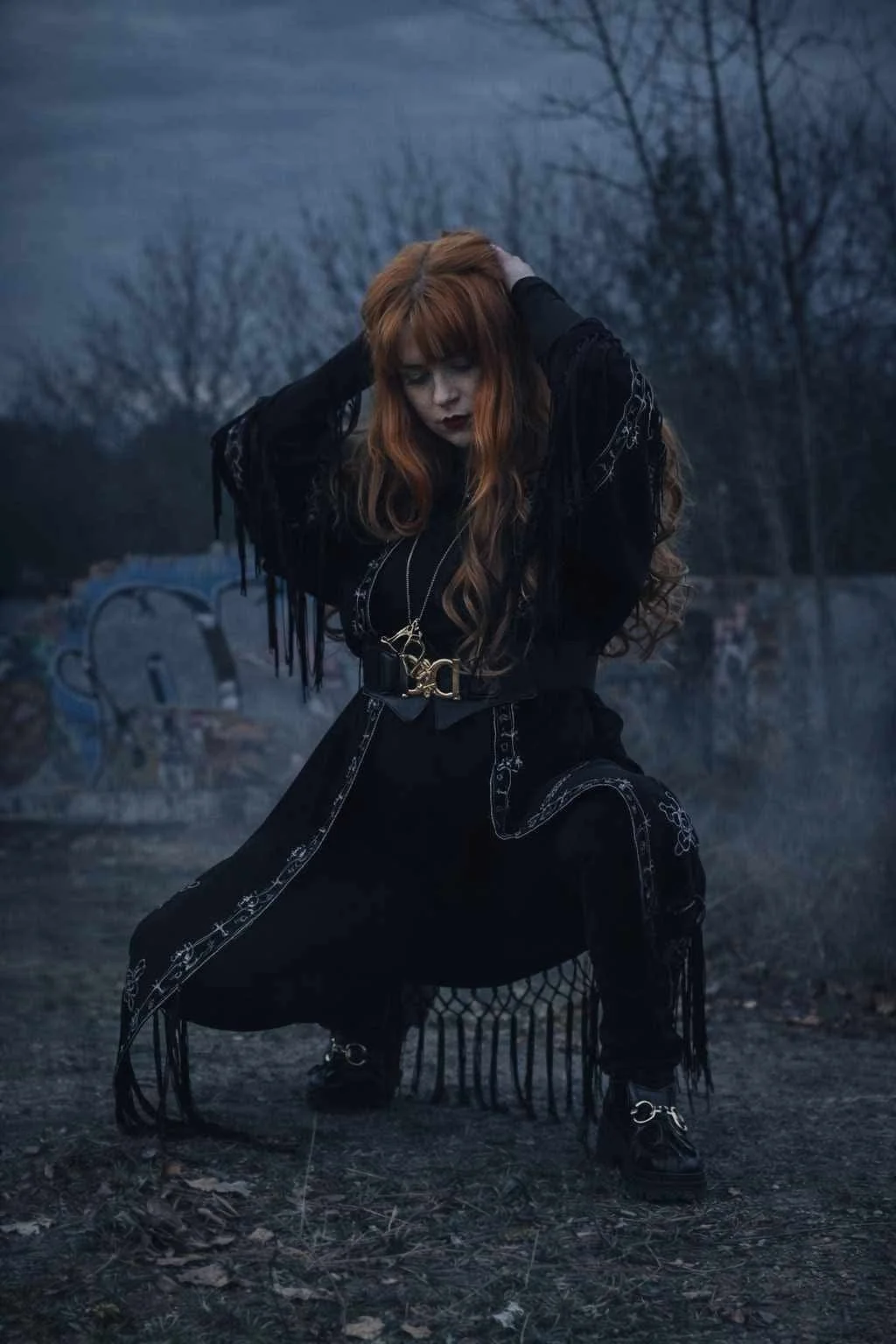 A woman with long, wavy, red hair, dressed in black gothic style clothing, is kneeling outdoors near a body of water during dusk, with trees and a bridge in the background.