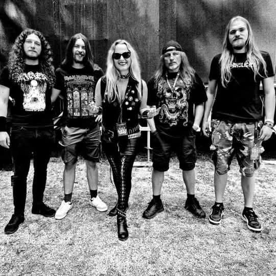 Group of five people standing outdoors in front of a dark wall, dressed in casual and edgy clothing, some with graphic t-shirts and one with sunglasses and a leather jacket.
