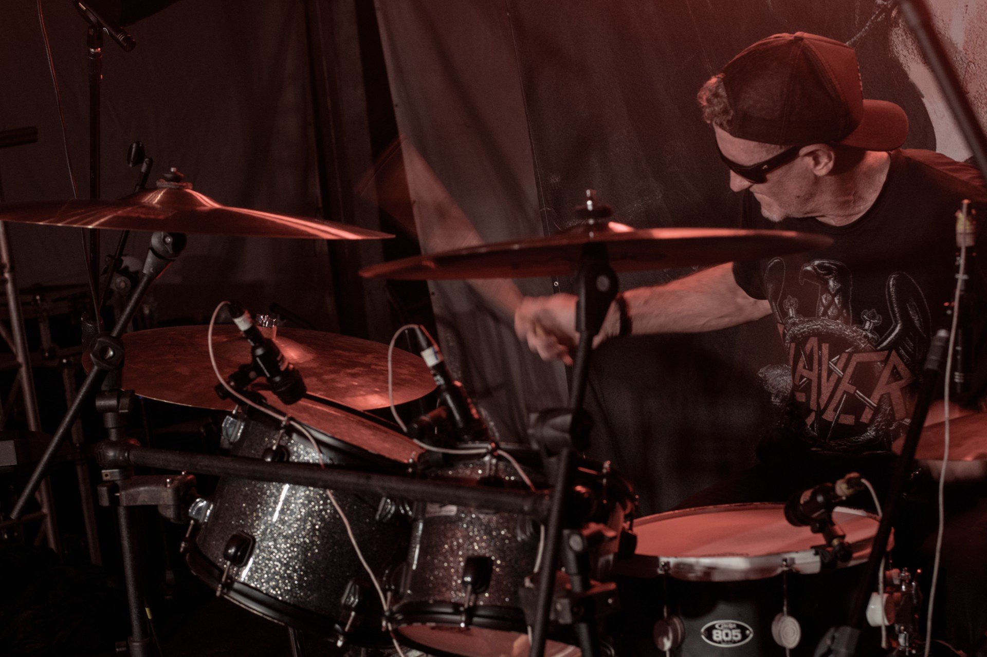 A man wearing a cap and sunglasses playing drums on stage under dim red lighting.