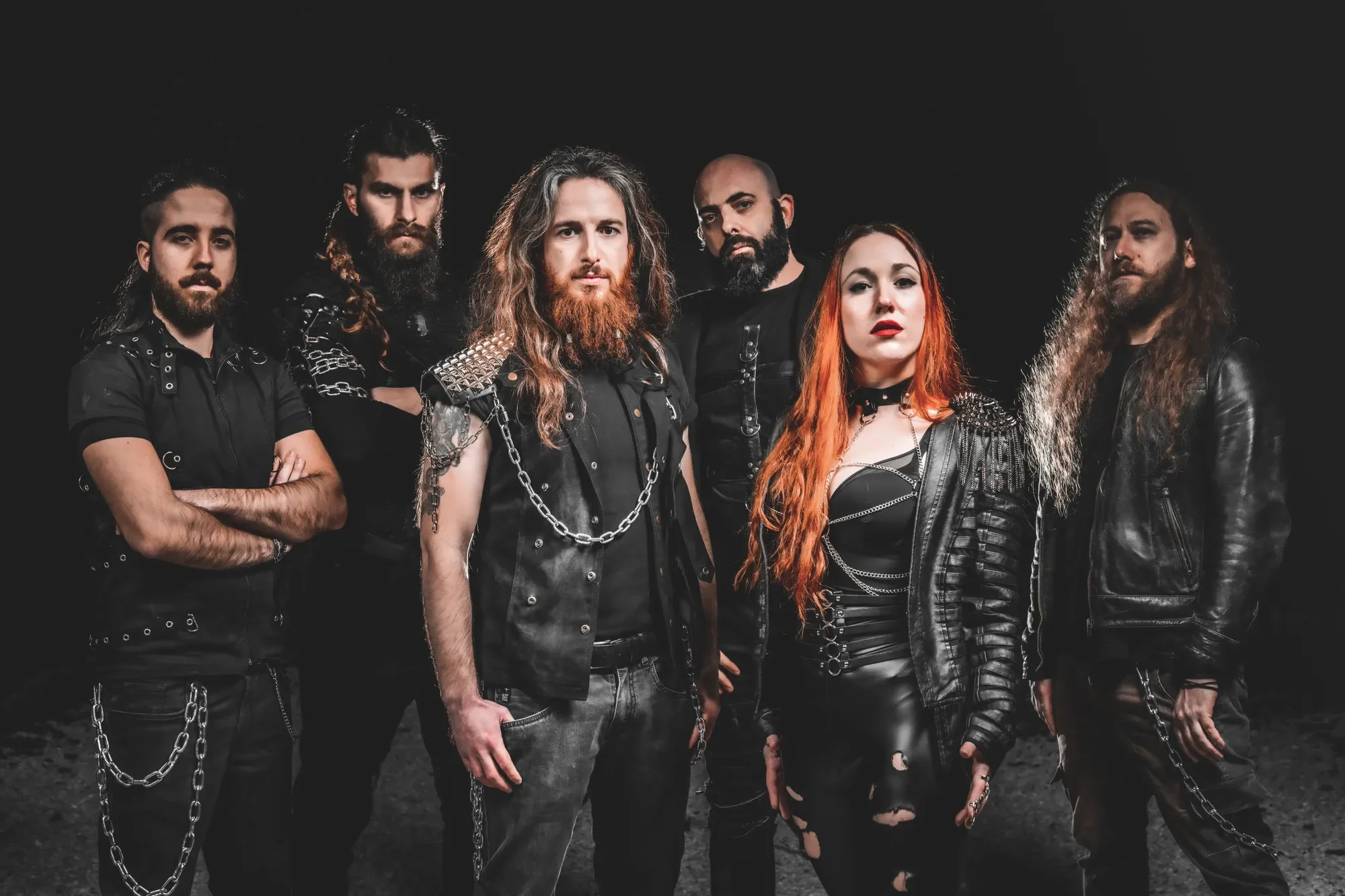 A group of six people in black leather clothing with chains, standing in front of a dark background, with serious expressions.