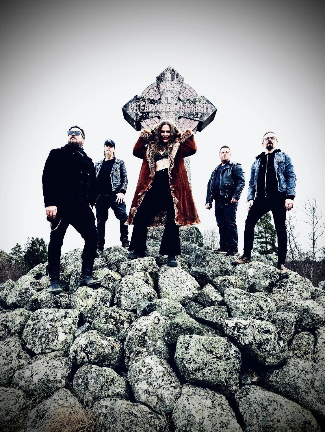 A group of five people standing on a pile of rocks outdoors around an old, weathered cross monument. The woman in the center is holding the top of the cross and wearing a fur-lined coat. The others are dressed in casual, dark clothing, and the background features a cloudy sky and sparse trees.