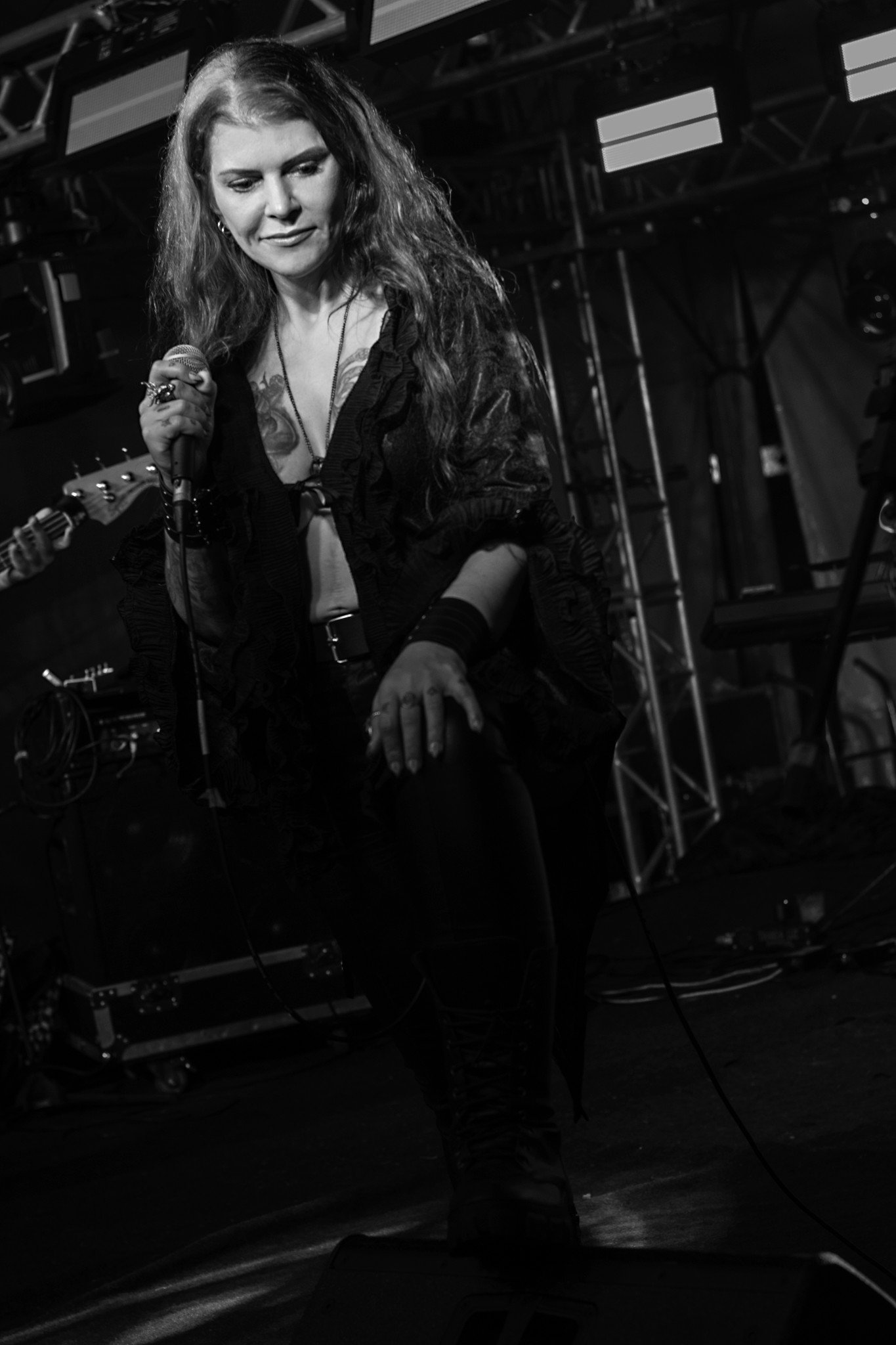 A black and white photo of a woman with long, wavy hair holding a microphone, standing on a stage with equipment and scaffolding in the background.