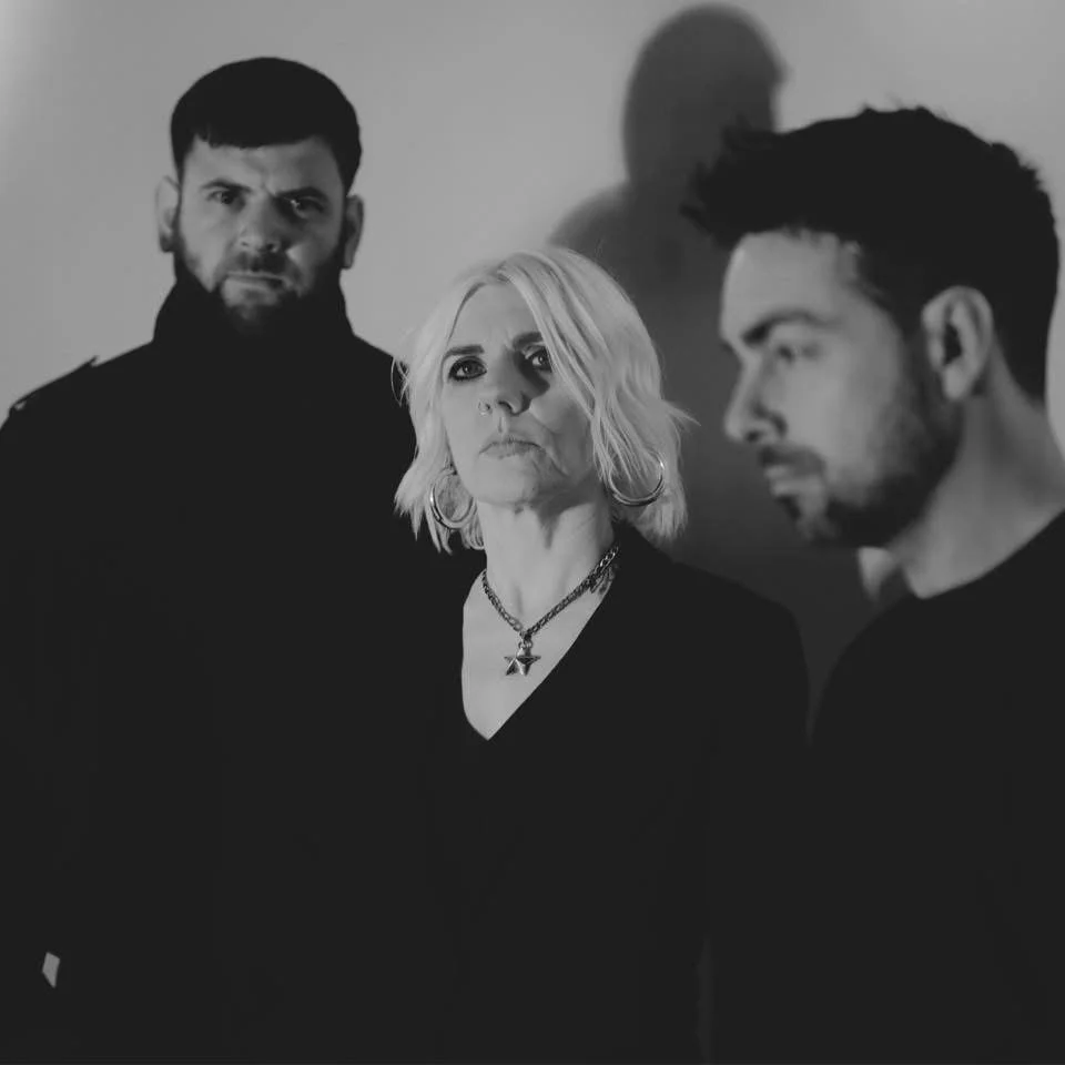 Black and white photo of three people, with a woman in the center, flanked by two men. The woman has short, blonde hair and wears a necklace and hoop earrings. The man on the left has dark hair and a beard, and the man on the right has dark hair and is looking to the side.