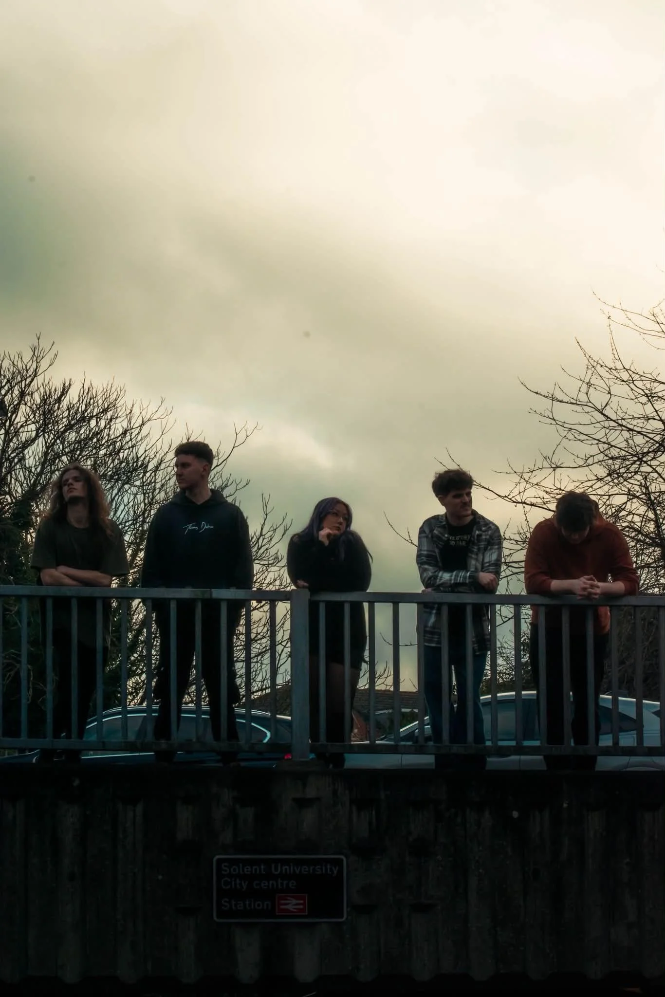 People standing on a bridge over a city street with leafless trees, cloudy sky, and a sign indicating Solent University, City Centre, and Station.