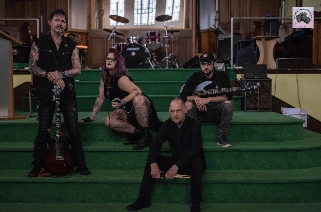 Four musicians sitting and standing on green steps inside a wood-paneled room with musical instruments in the background.