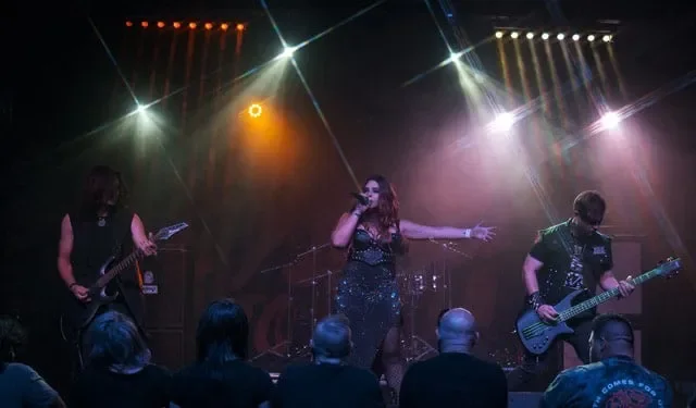 A band performs on stage with singer and two guitarists, colorful stage lights, and audience watching.