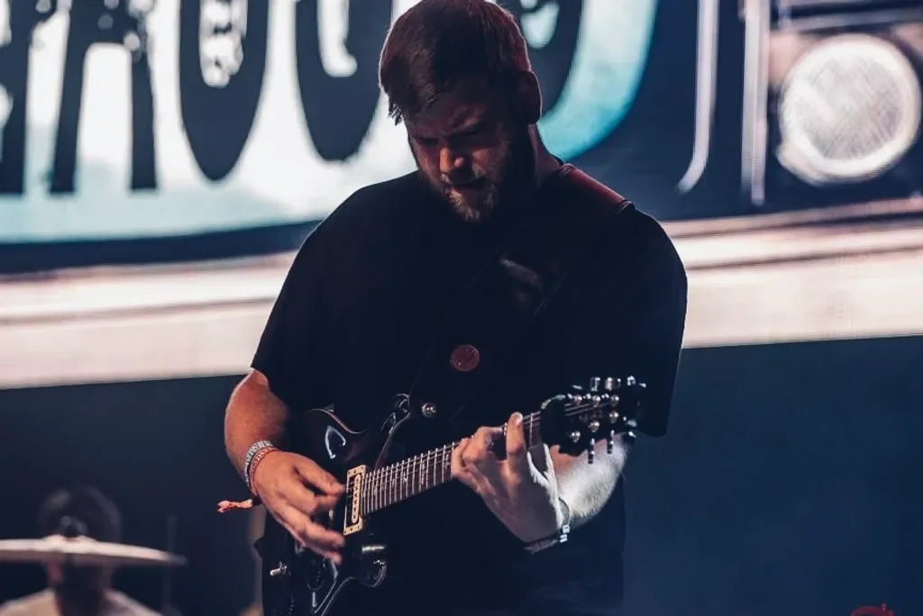 A man playing an electric guitar on stage with a dark background and a large screen displaying abstract graphics.