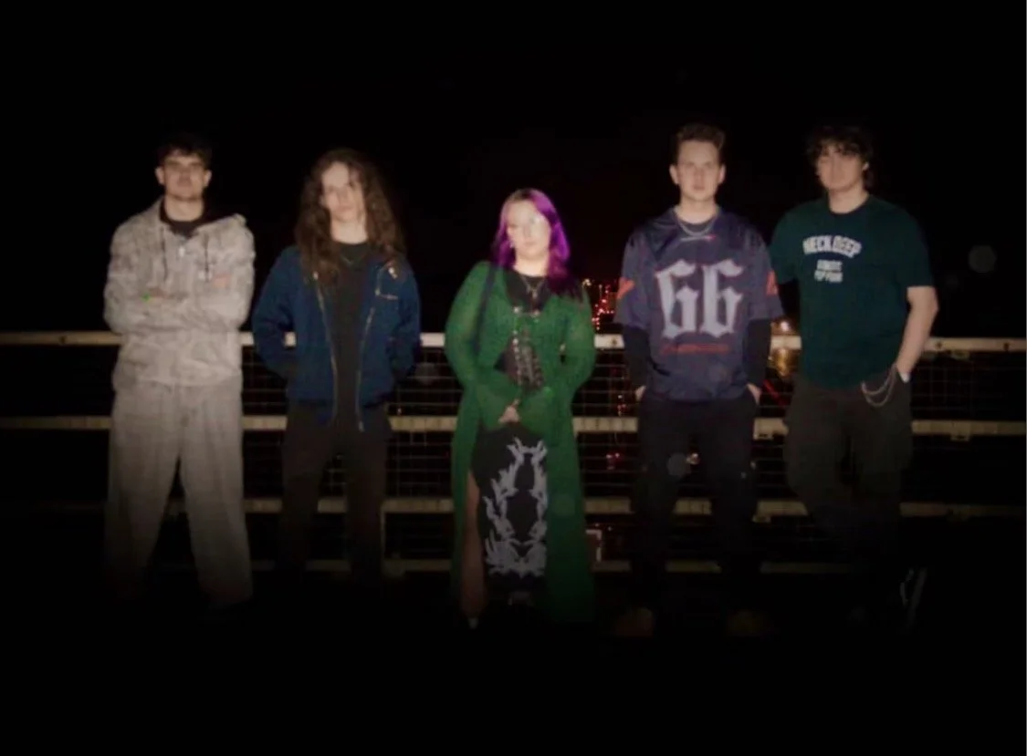 Five people standing outdoors at night behind a metal railing, with dark sky and city lights in the background.